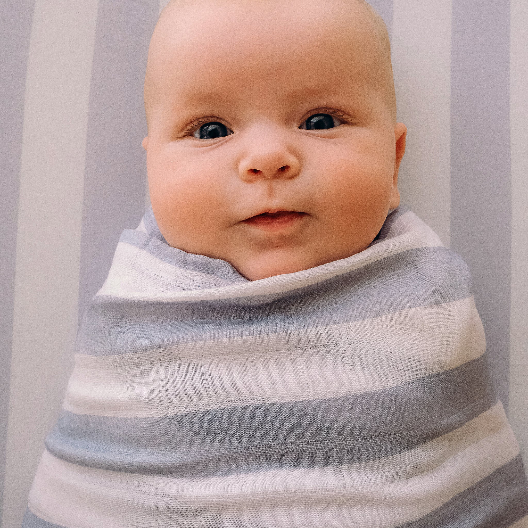 A baby with wide blue eyes and a slight smile, wrapped in a white and gray striped blanket, rests on the FOX & FALLOW - Cloud Stripe Organic Fitted Cot Sheet, featuring GOTS certified organic cotton.