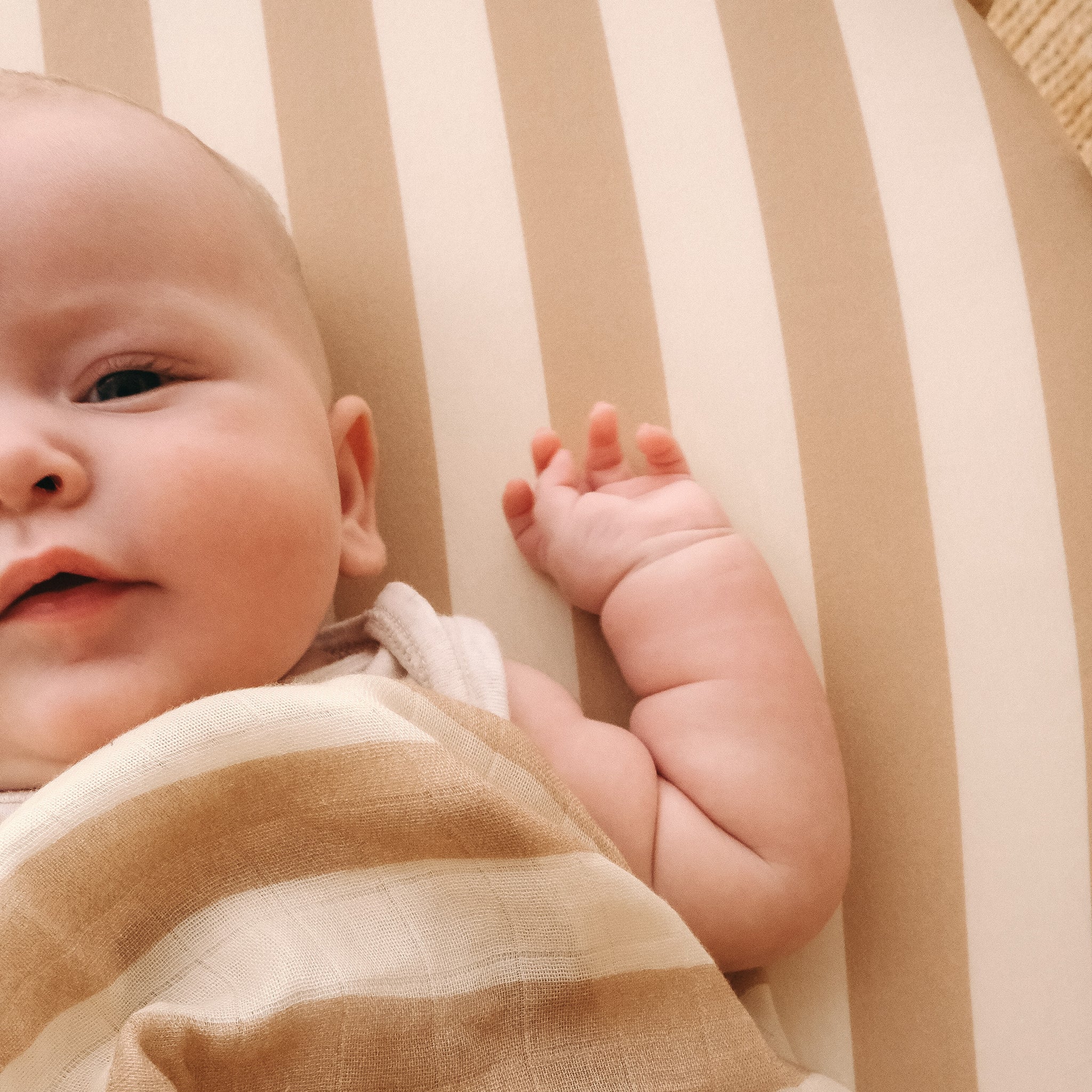 A baby lies on the Fox & Fallow - Coffee Stripe Organic Fitted Bassinet Sheet/Change Pad Cover by FOX & FALLOW, gazing at the camera with a slight smile and one arm raised, partially wrapped in the cozy, GOTS-certified striped fabric.