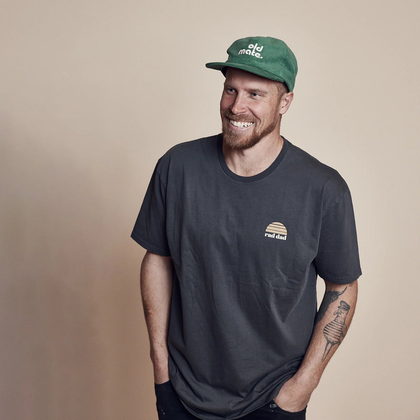 A smiling man with a beard wears the Banabae - Rad Dad Organic Cotton Tee in washed black by BANABAE, paired with a green cap. Perfect Father’s Day or Dad gift, he stands against a plain beige background with hands in pockets.