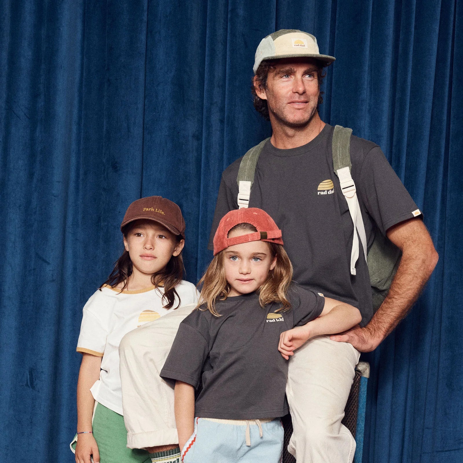 A man in a BANABAE "Rad Dad" Organic Cotton Tee (Washed Black) stands behind two kids, all in casual outfits and caps, posing by a blue curtain—a great Father's Day snapshot.