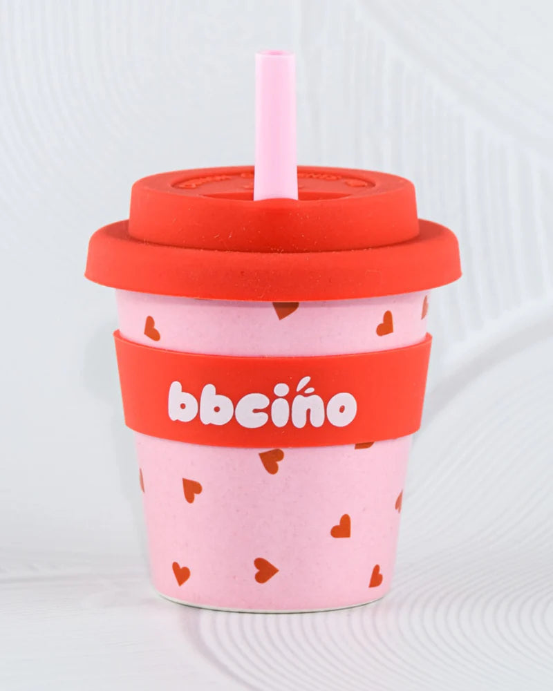 The BBCINO - Reusable Babycino Cup 120ml Sweetheart features a pink cup with red hearts, a red lid, and pink straw. Made from sustainably farmed bamboo, it has a red band with "bbcino" in white on a textured light background.