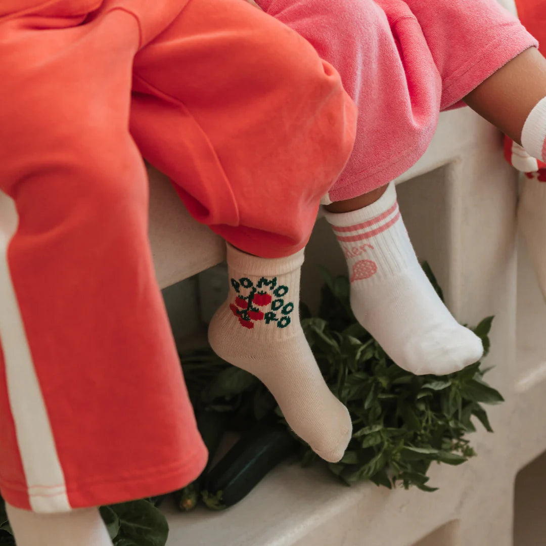 Two children sit side by side, one in coral pants and GOLDEN CHILDREN Golden Children - Pomodoro Socks Dough with tomato and “POMODORO” print and reinforced toe caps, the other in pink pants and white socks. Green vegetables are placed below them.