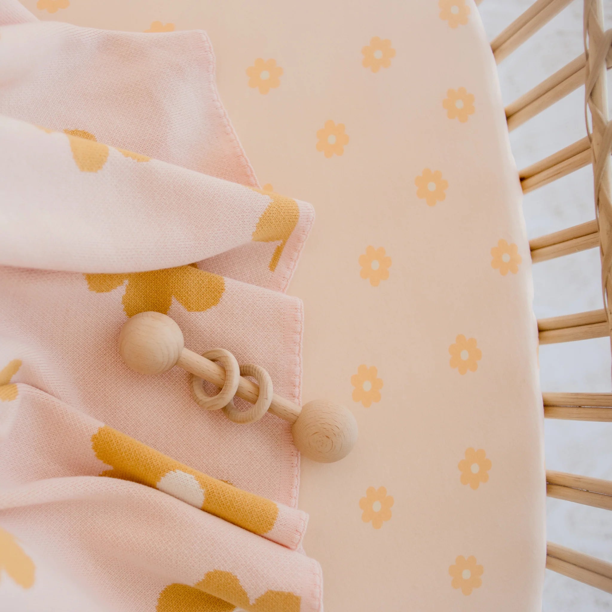 A close-up of the Fox & Fallow - Daisy Chain Organic Fitted Bassinet Sheet/Change Pad Cover by FOX & FALLOW, featuring a peach-colored GOTS certified fabric with small yellow flowers, alongside a floral blanket and wooden baby rattle on a woven crib.