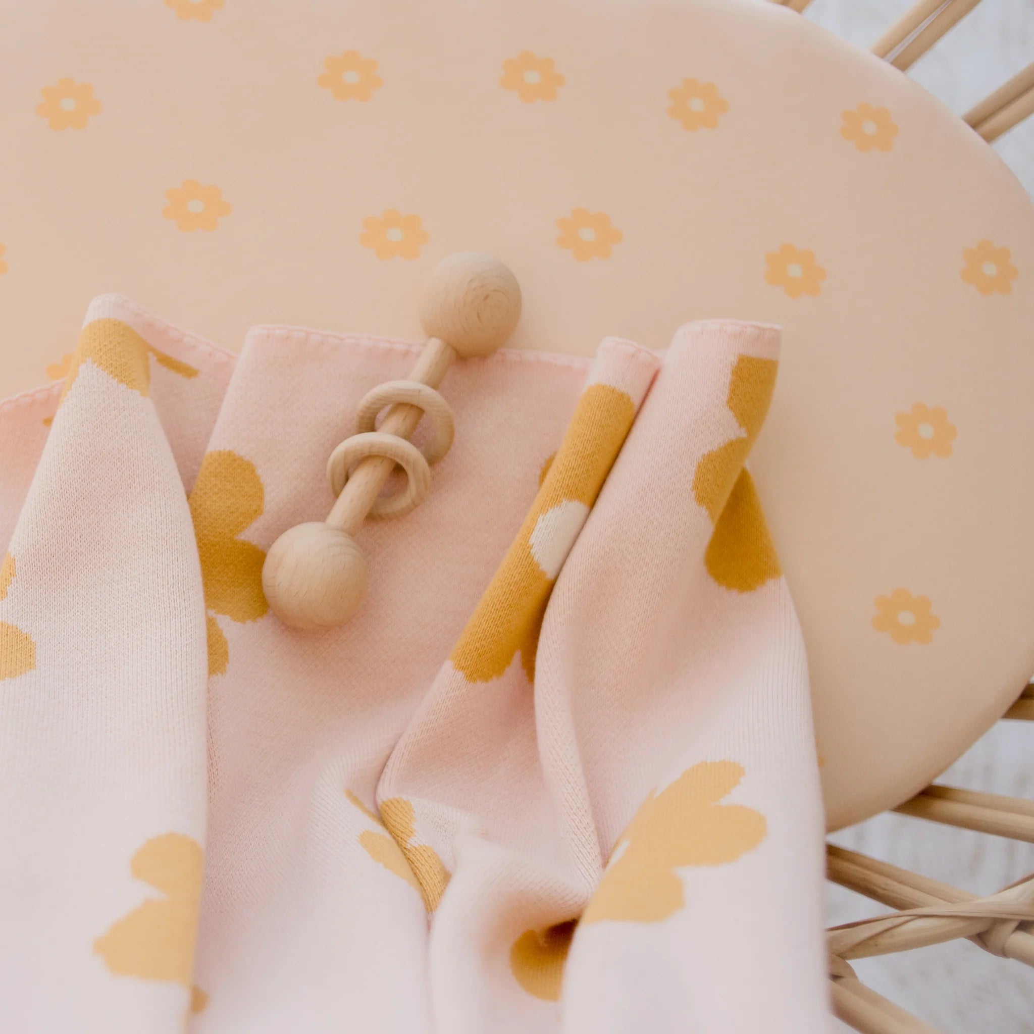 A wooden baby rattle sits on a soft pink blanket with yellow flowers, atop a peach chair adorned with matching florals and the FOX & FALLOW Daisy Chain Organic Fitted Bassinet Sheet/Change Pad Cover.
