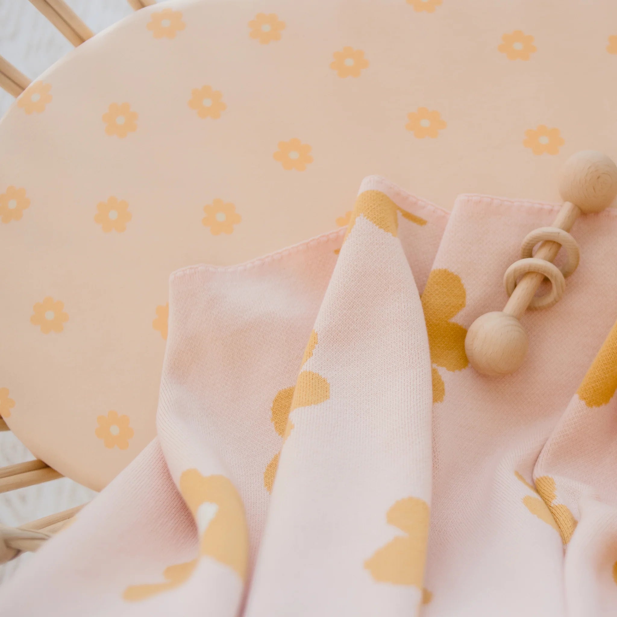 A soft pink blanket with yellow florals rests on a matching chair beside a wooden baby rattle—perfectly complementing our FOX & FALLOW Daisy Chain Organic Fitted Bassinet Sheet/Change Pad Cover.