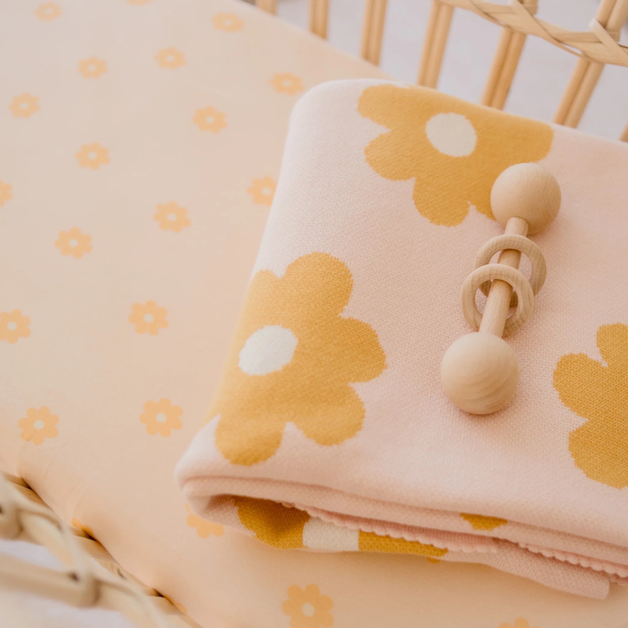 A folded pink blanket with yellow and white flowers and a wooden rattle rest on the Fox & Fallow - Daisy Chain Organic Fitted Bassinet Sheet/Change Pad Cover by FOX & FALLOW in a wicker crib.