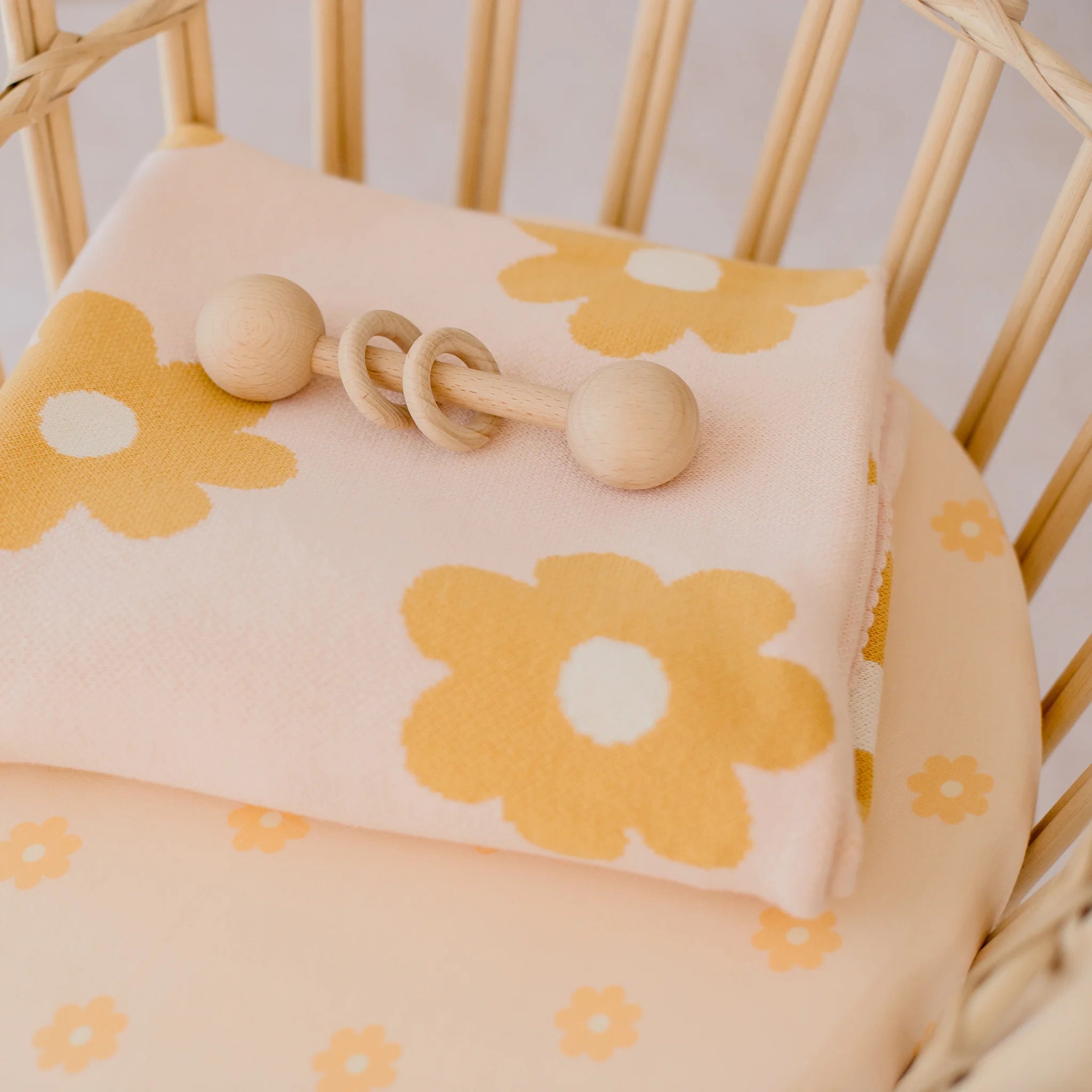 A soft baby blanket with yellow flower patterns rests on a cushioned chair, topped with a wooden rattle. Pair this cozy setting with the FOX & FALLOW Daisy Chain Organic Fitted Bassinet Sheet/Change Pad Cover for added comfort.