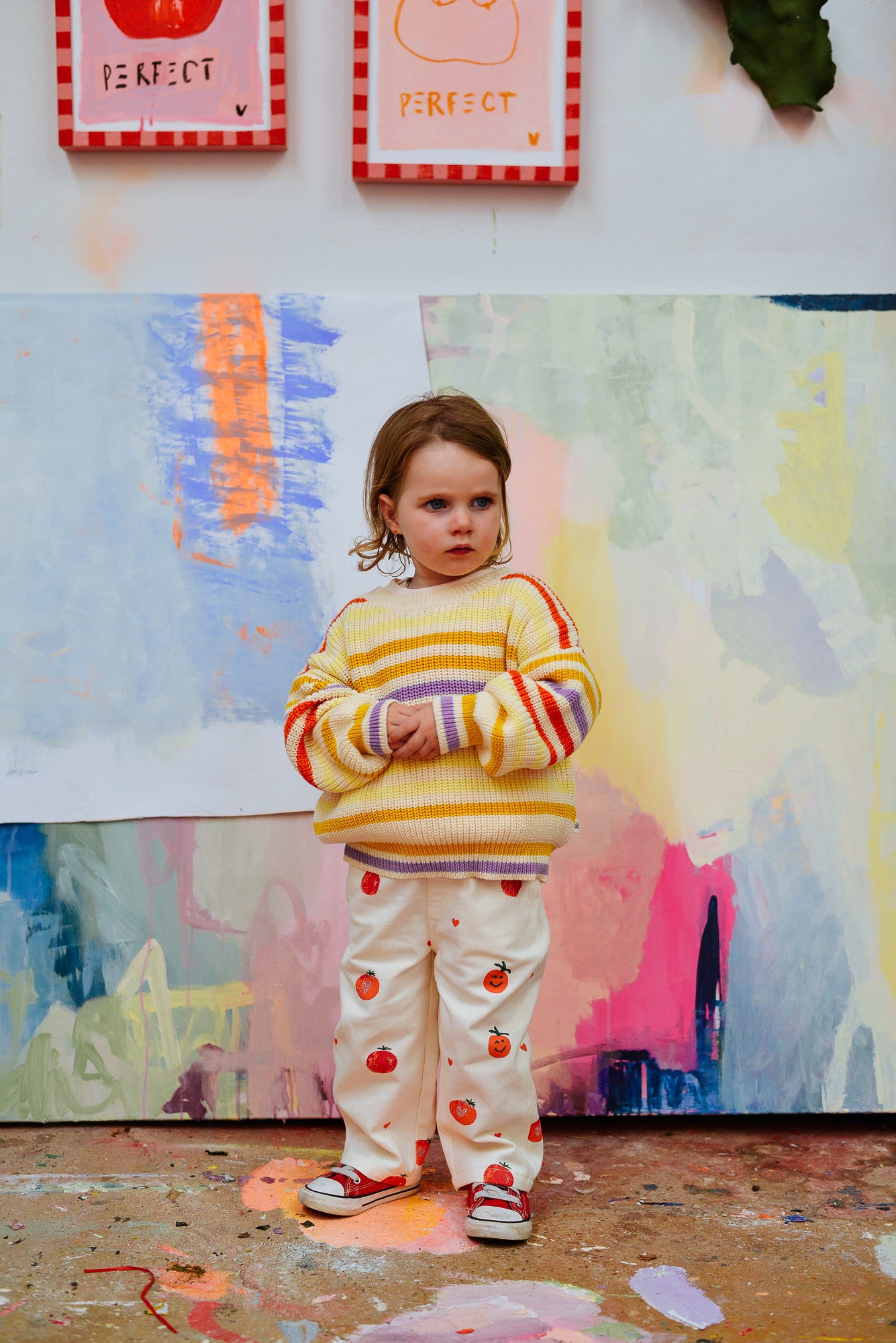 A young child stands indoors in the SUNDAY SIBLINGS Sunday Siblings - Art Club Knit sweater, white pants with red fruit prints, and red shoes, surrounded by abstract pastel artworks.