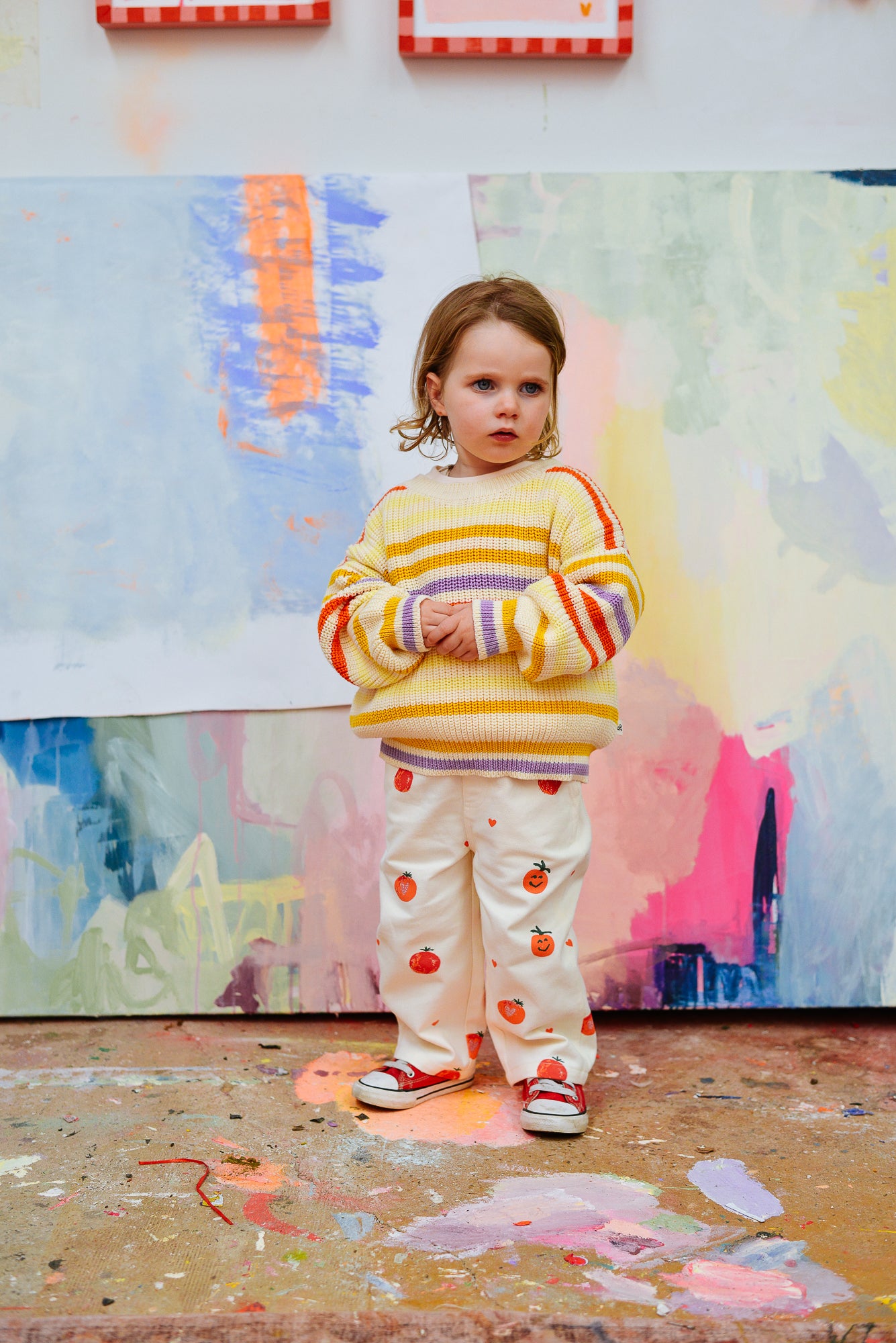 A young child stands indoors on a paint-splattered floor wearing the SUNDAY SIBLINGS Sunday Siblings - Art Club Knit sweater. Colorful abstract paintings hang and rest against the wall behind them.