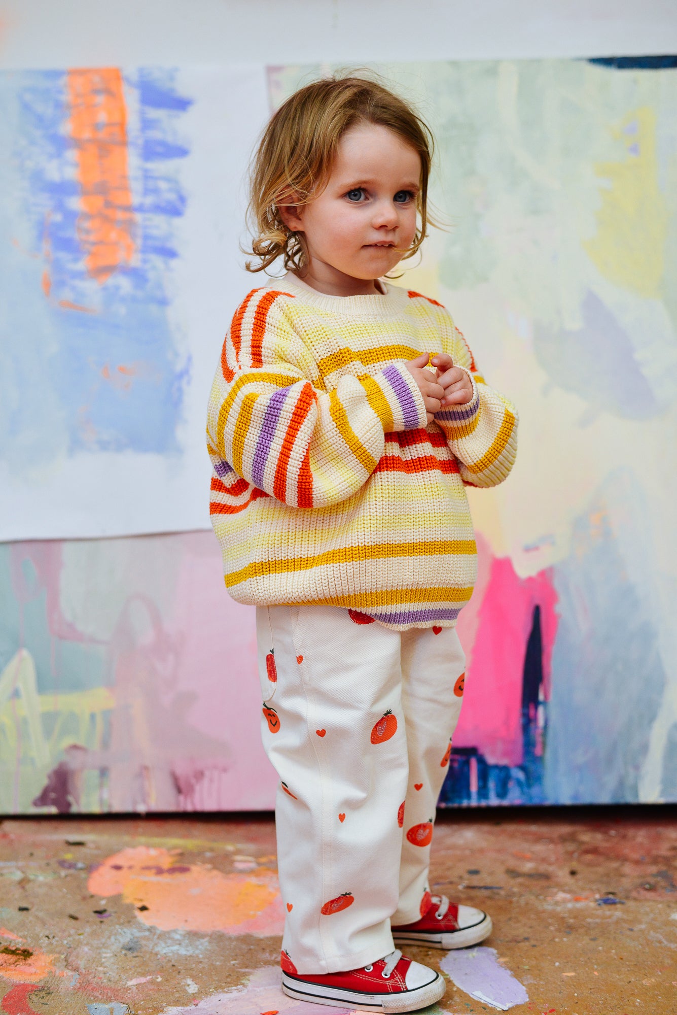 A young child in a yellow and orange striped Sunday Siblings - Art Club Knit sweater by SUNDAY SIBLINGS, white pants with red patterns, and red sneakers stands indoors against a vibrant, abstract painted backdrop.
