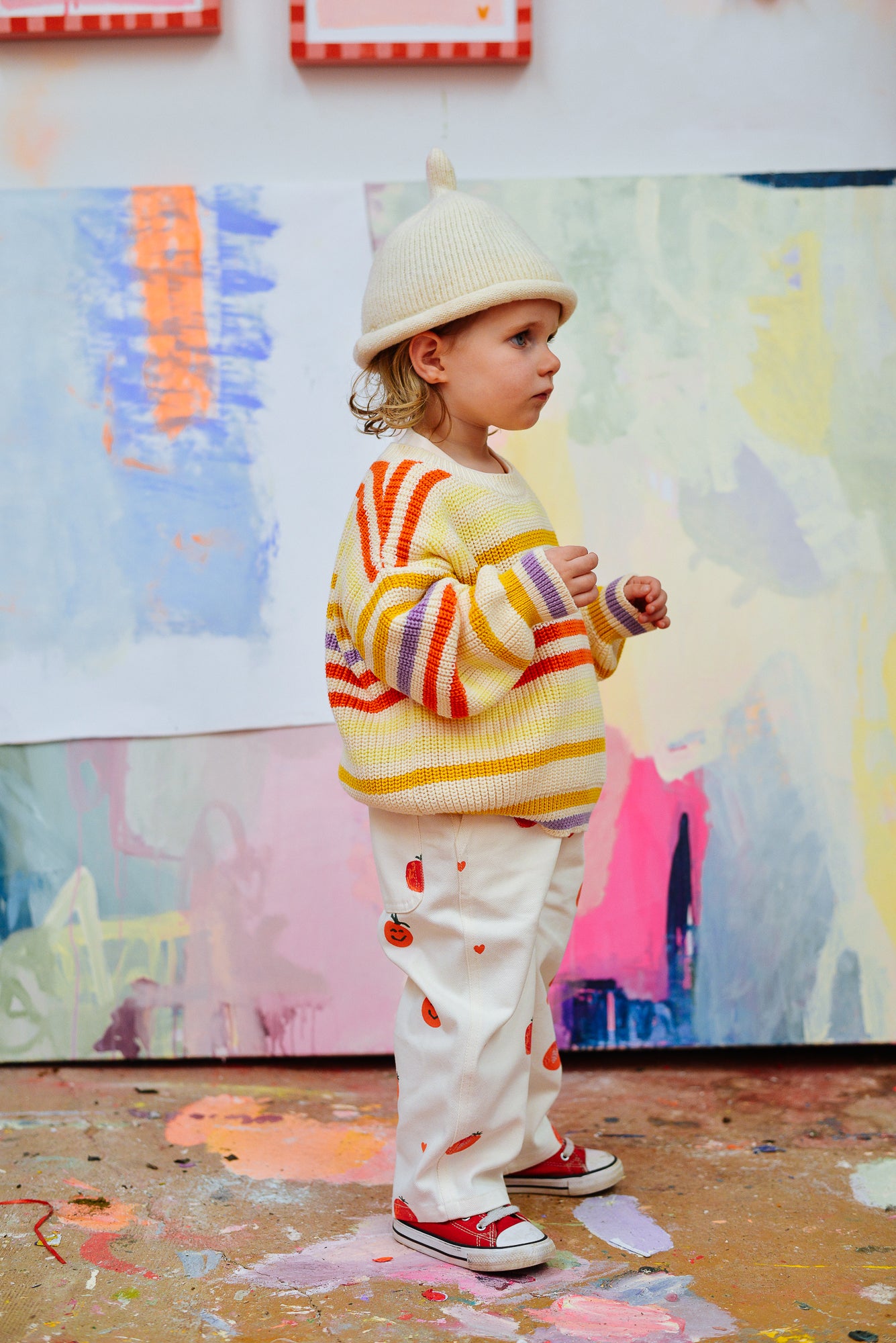 A toddler models the PRE-ORDER Sunday Siblings - Art Club Knit sweater by SUNDAY SIBLINGS, paired with a cream knit beanie, red sneakers, and patterned white pants, standing indoors against a pastel painted backdrop.