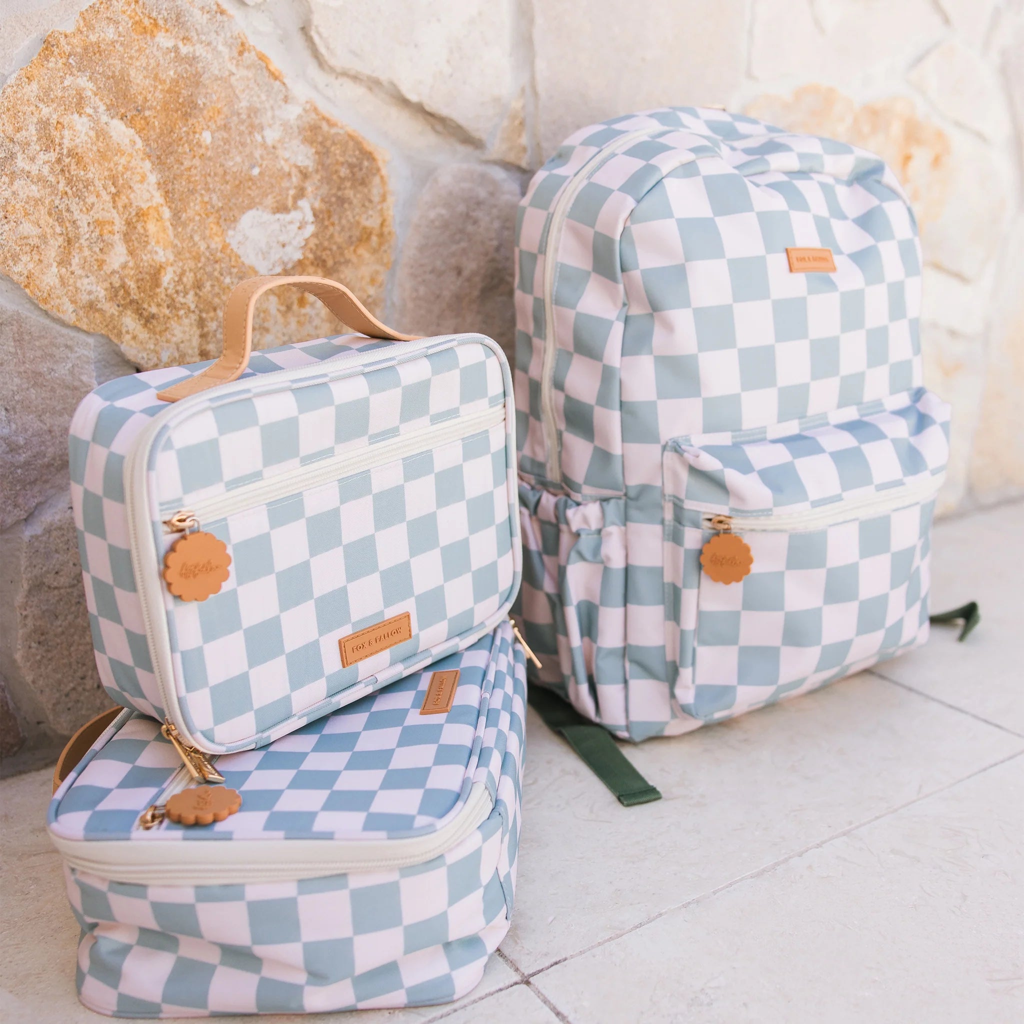 The Fox & Fallow - Sage Check Large Lunch Bag by FOX & FALLOW, featuring a light blue and white checkered pattern, sits on a light stone floor against a stone wall.