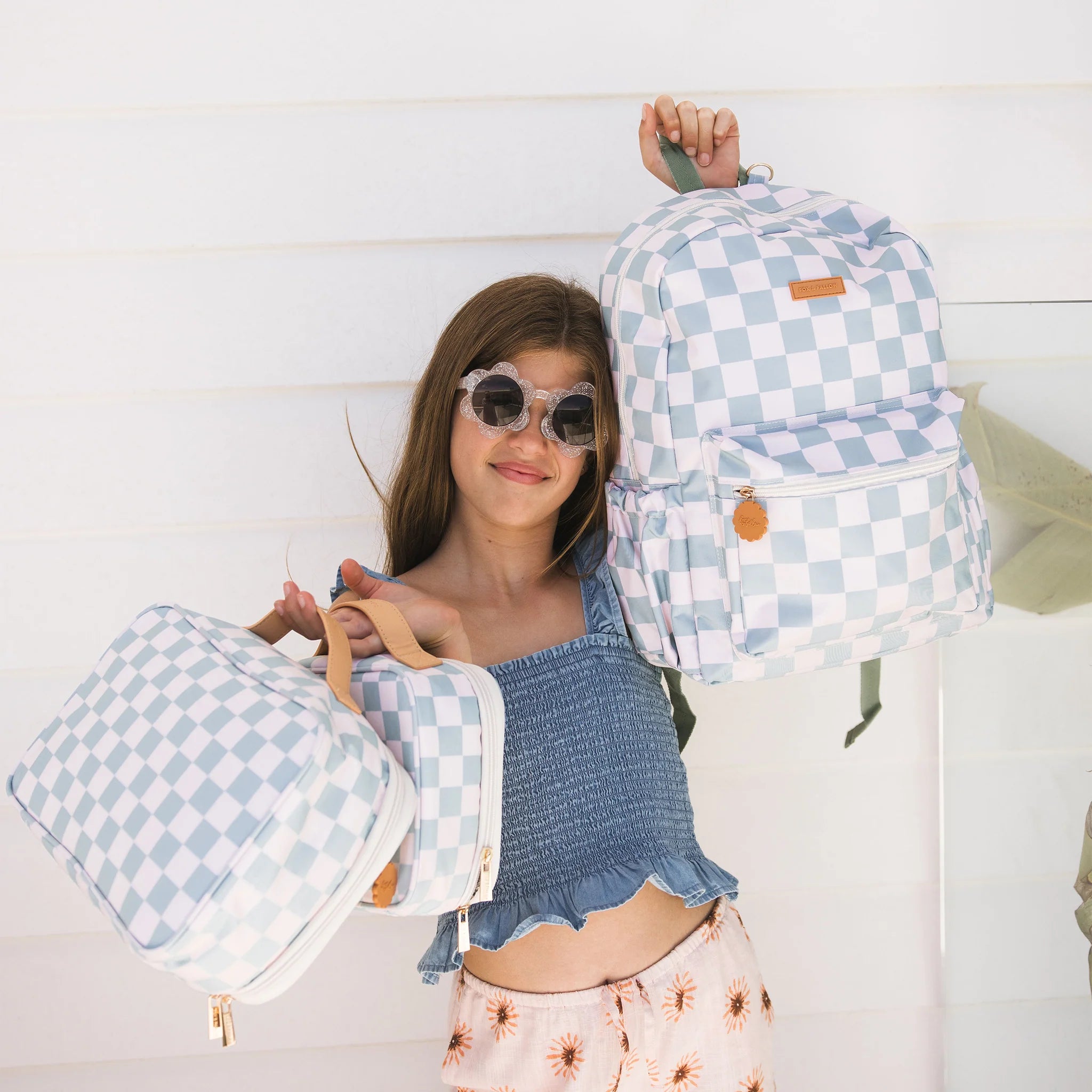 A girl in sunglasses and a blue top smiles while holding the FOX & FALLOW Sage Check Large Lunch Bag and a matching backpack, standing in front of a white wall.