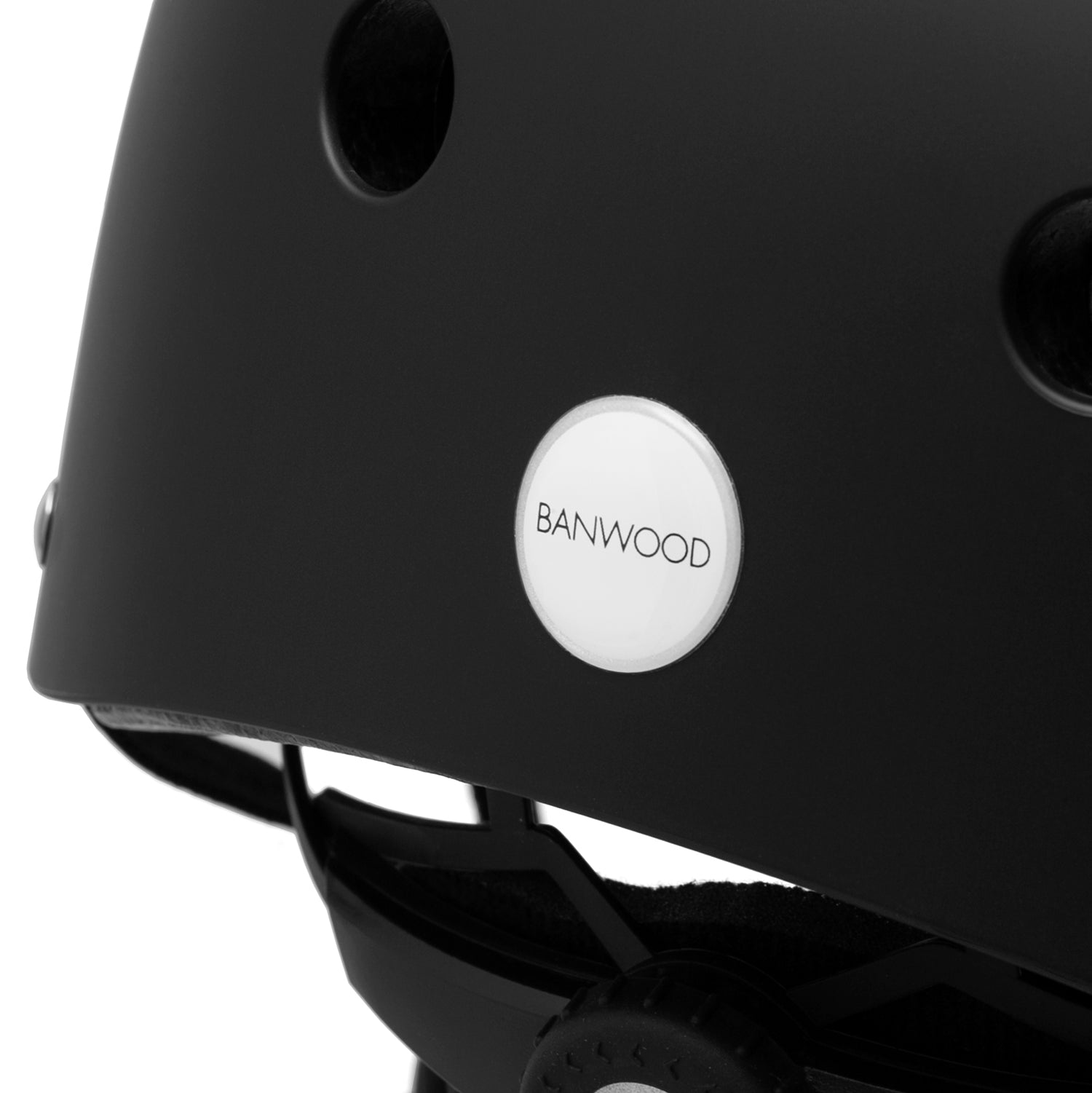 A close-up of the Banwood - Classic Helmet Black - S shows a round white "BANWOOD" logo on its side. This kids' bike helmet features ventilation holes and adjustable straps for a secure, comfortable fit, ensuring optimal protection during every ride.