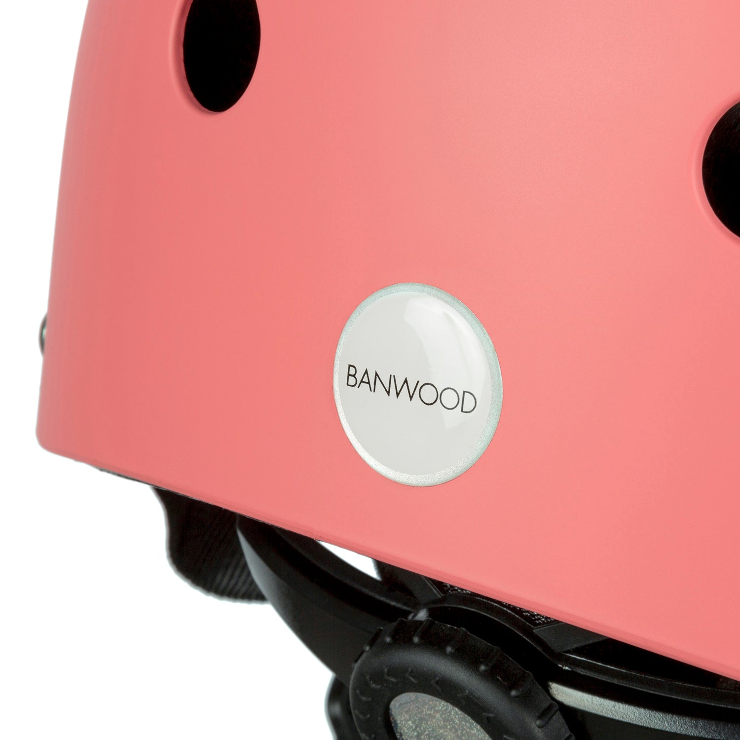 Close-up of a coral Banwood Classic Helmet - S, showing the "BANWOOD" name on a round white label. The helmet includes ventilation holes and an adjustable black strap for optimal children's protection.