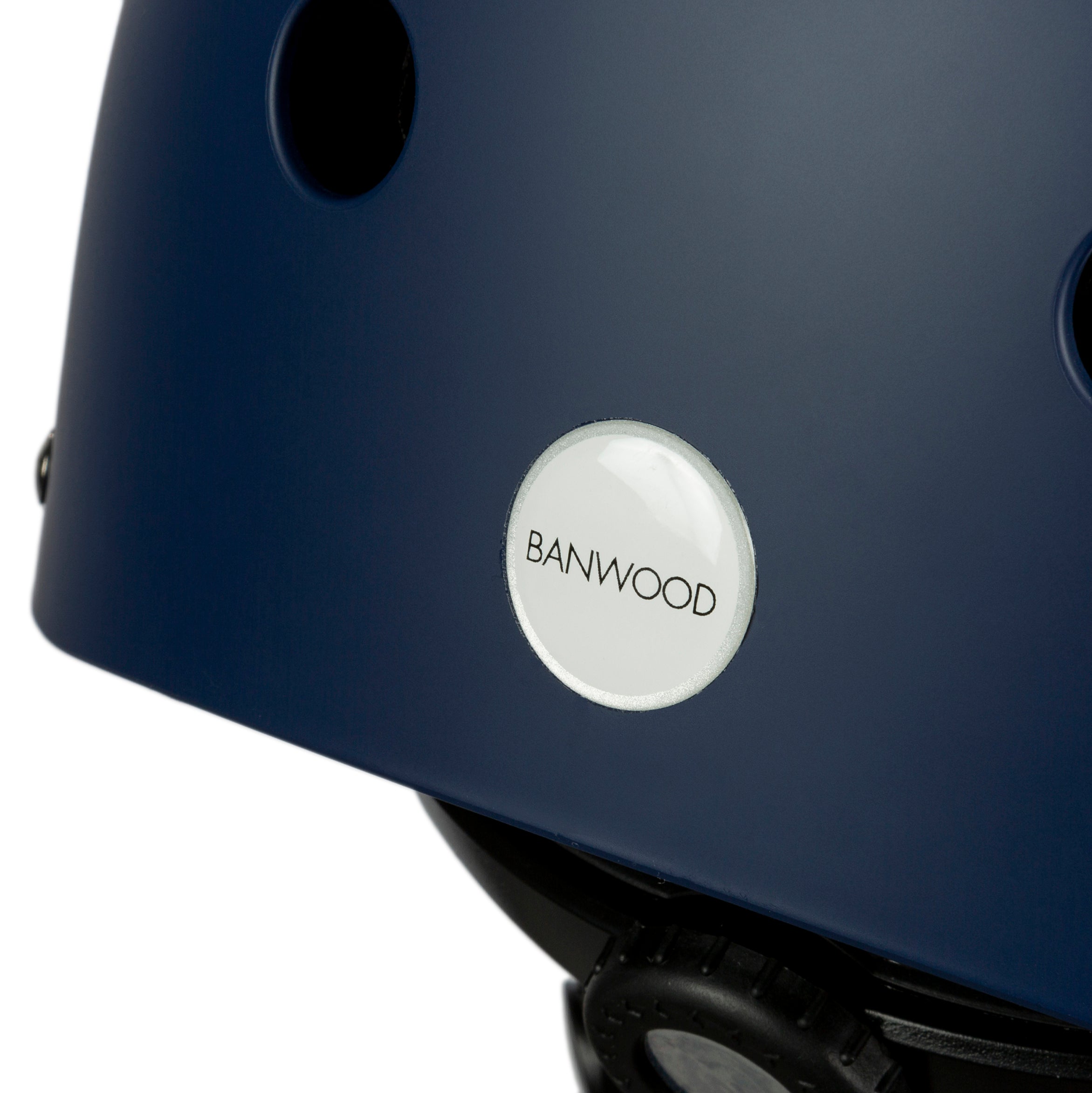 Close-up of the Banwood - Classic Helmet Navy - S, featuring ventilation holes and a white circular "BANWOOD" sticker. The helmet strap and buckle ensure security with its dial fit system, offering kids protection.