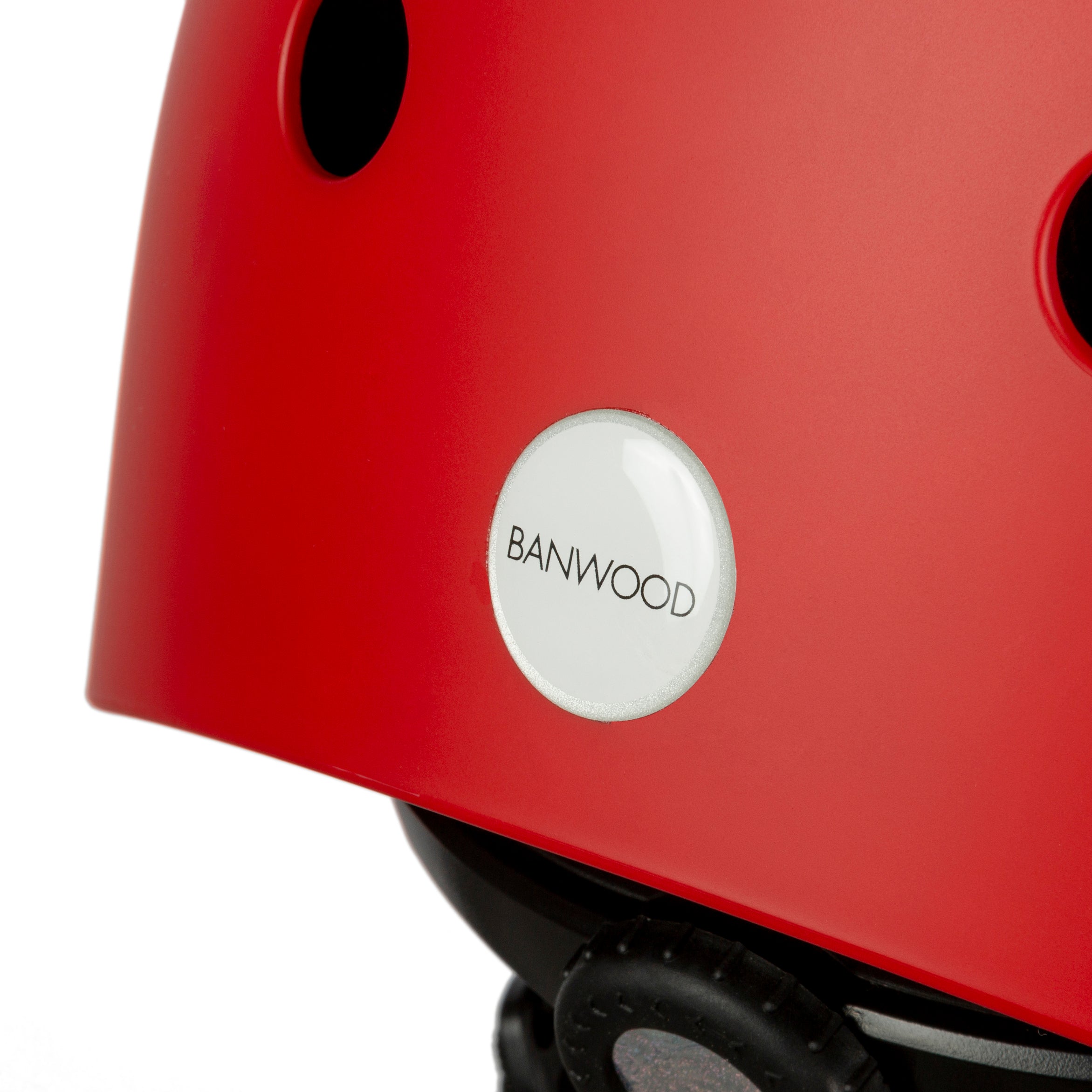 Close-up of the “Banwood - Classic Helmet Red - XS,” featuring black ventilation holes and the BANWOOD logo on a white circle. This children's bike helmet includes an adjustable strap with a rear buckle for a secure fit.