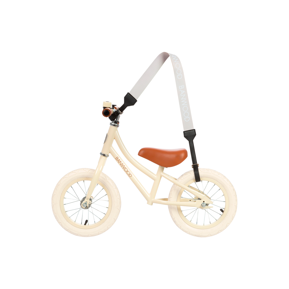 A beige child's balance bike with white tires, a brown seat, and the Banwood Carry Strap in Cream for easy transport. It includes a small bell on the handlebar, offering style and functionality for young riders.