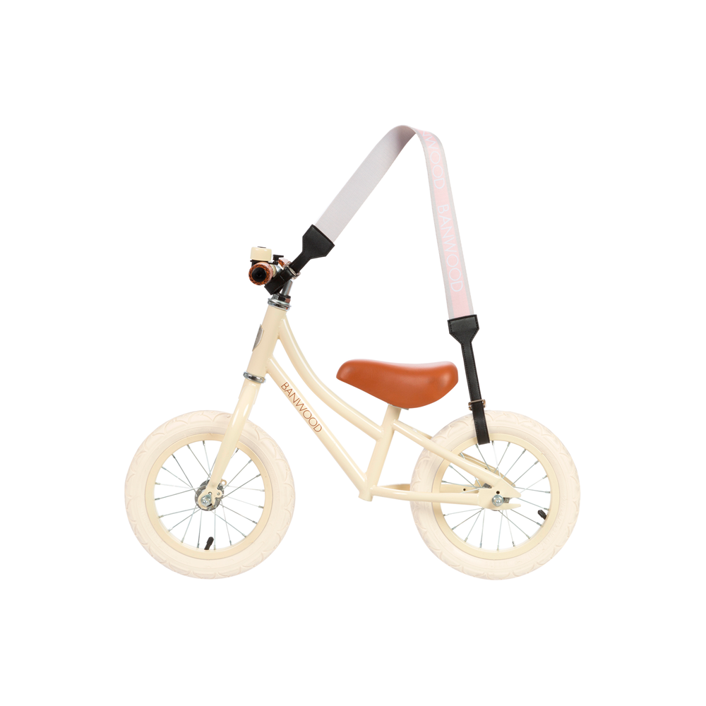 A beige balance bike with white tires and a brown vegan leather seat includes the Banwood Carry Strap Pink, allowing easy transport by attaching to the handlebars and back of the seat.