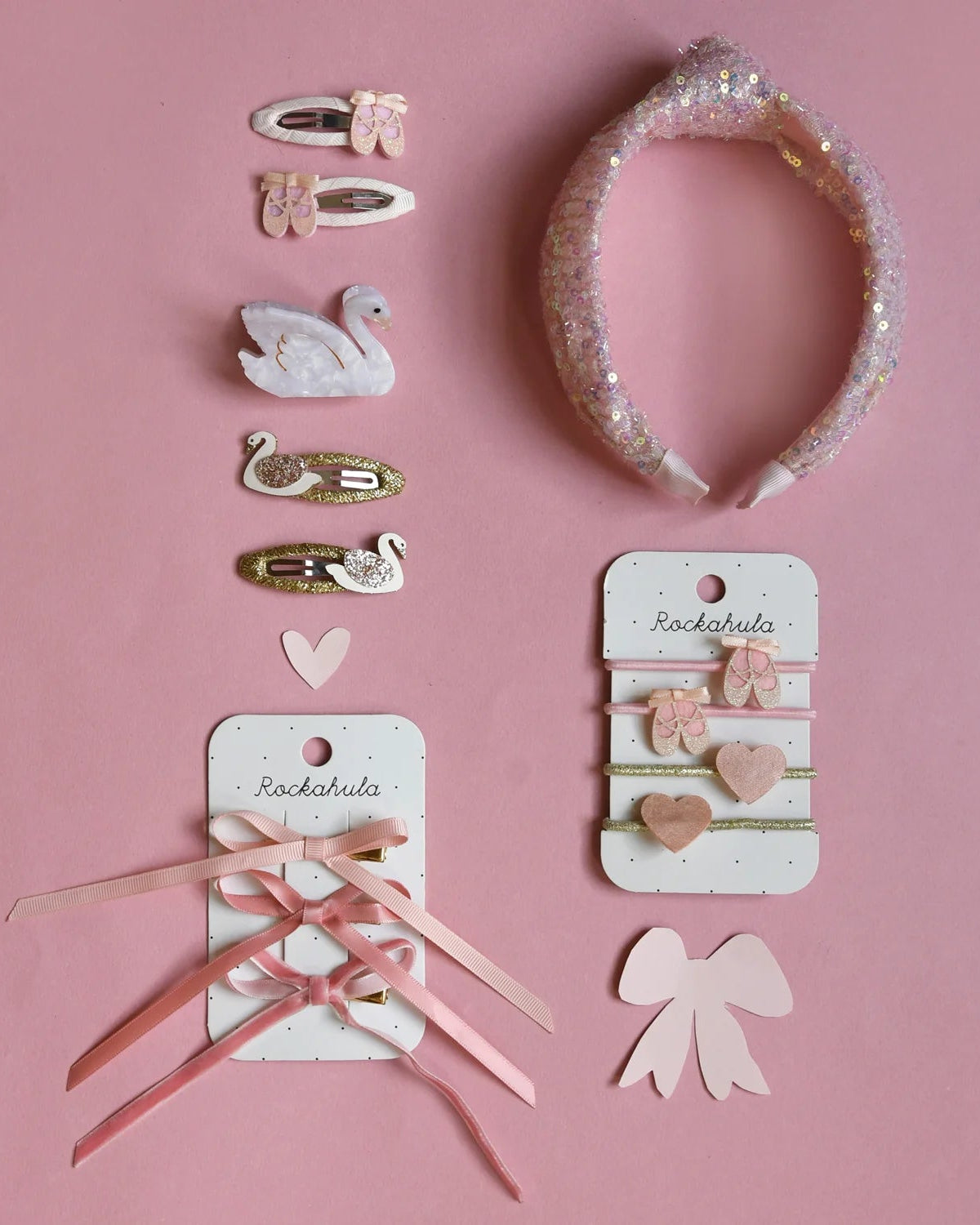 Rockahula's Ballet Shoes Ponies by ROCKAHULA are pink and white hair accessories, including barrettes with bows and swans, a glittery headband, ribbon ties, and cards with hair clips—perfectly arranged on a pink background.