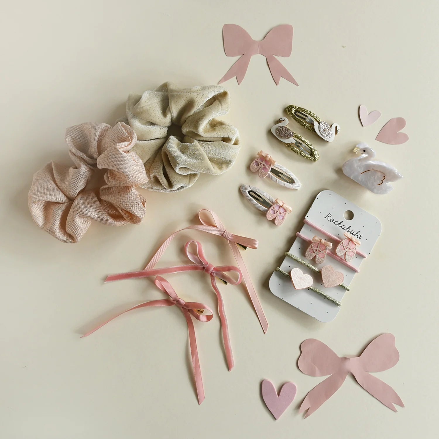 The ROCKAHULA 'Ballet Shoes Ponies' hair accessories set features pastel scrunchies, pink glitter clips with floral and animal motifs, and ribbon bows, all arranged on a light background with paper hearts and bows.