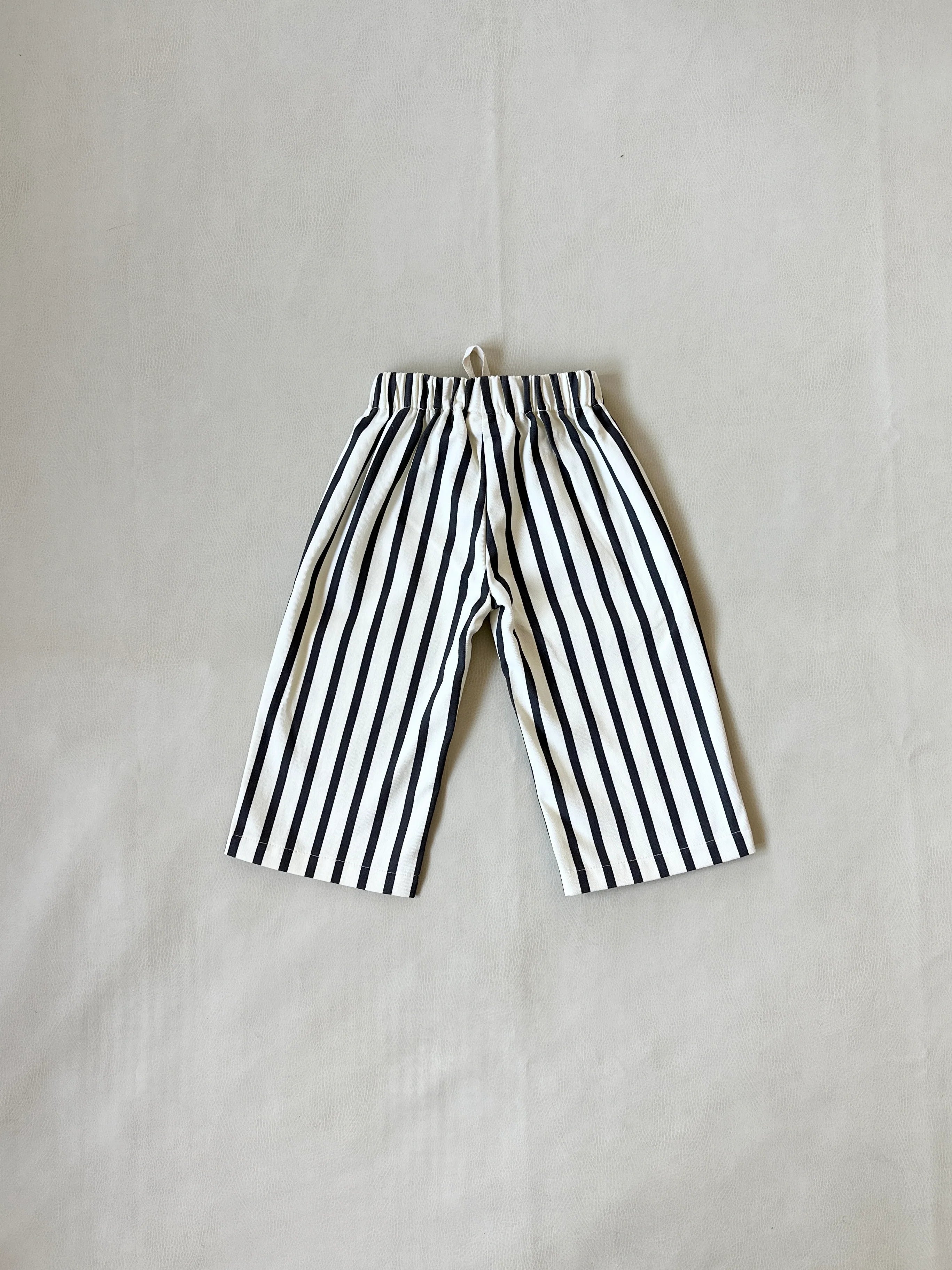 A pair of TINY TROVE Billie Stripe Cotton Pants Cream/Cocoa Stripe for kids with vertical stripes, laid flat on a light-colored surface.
