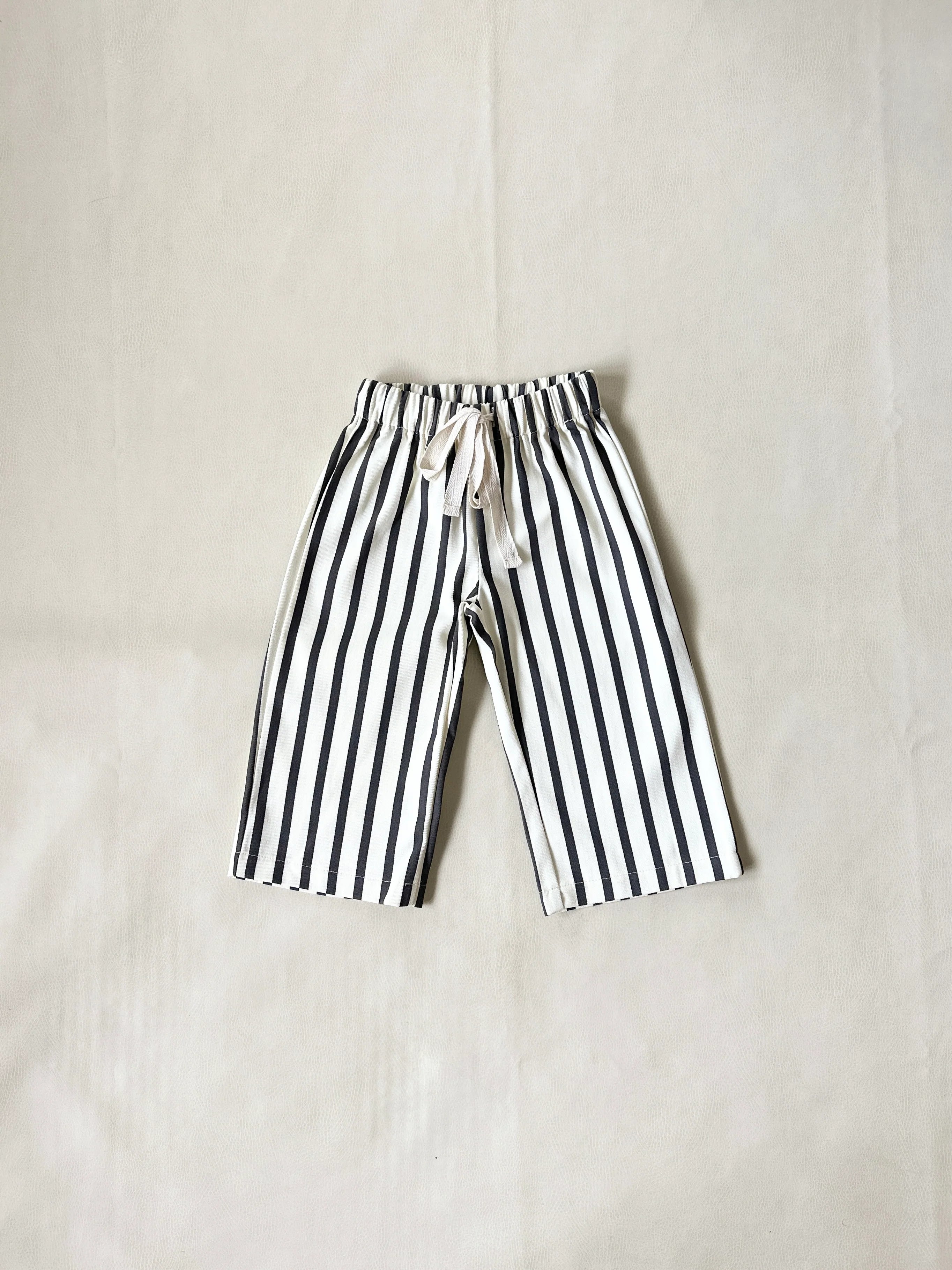 A pair of TINY TROVE Billie Stripe Cotton Pants in Cream/Cocoa Stripe, knee-length with vertical stripes and an elastic waist with white drawstring, laid flat on a light beige surface.