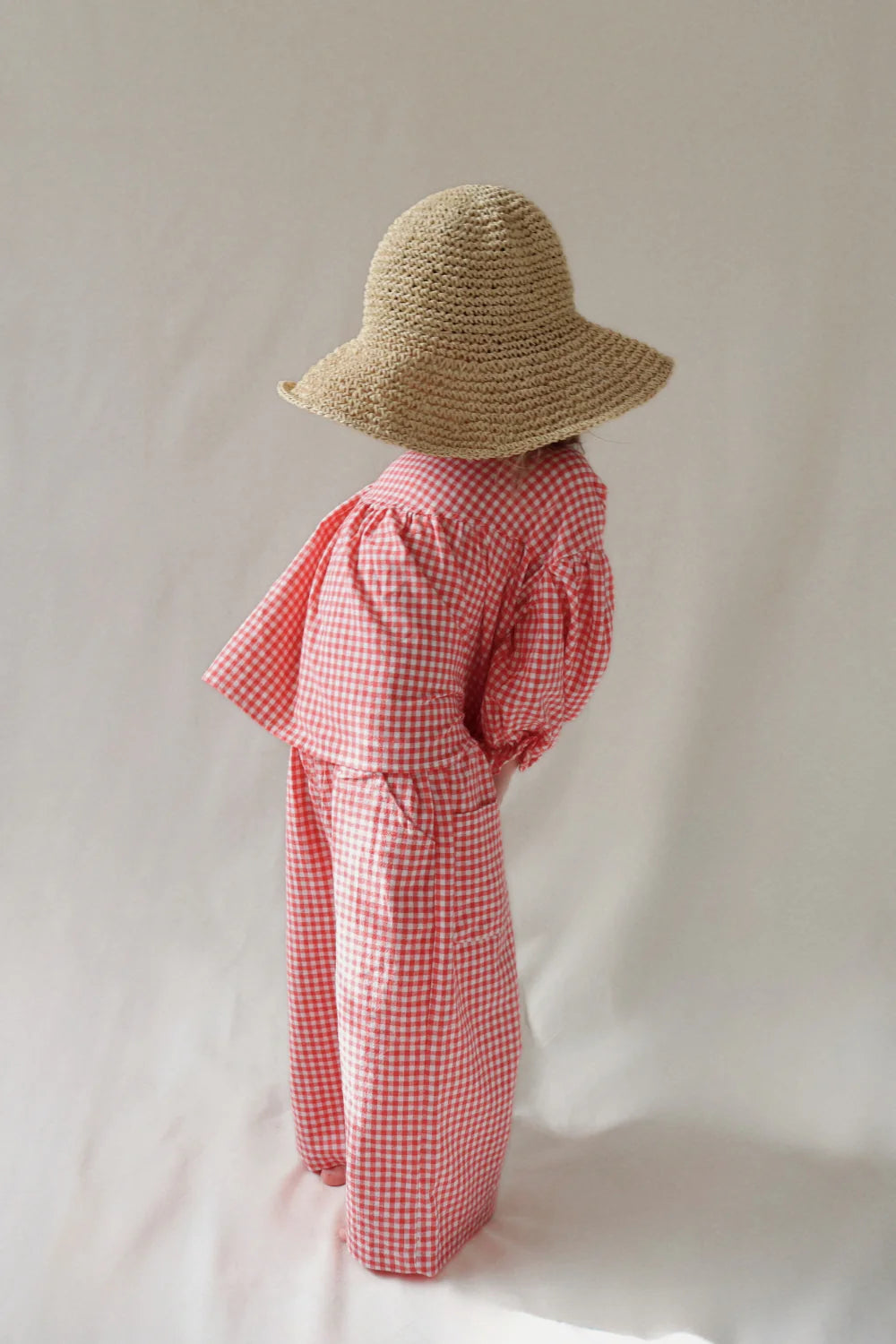 A young child faces away, wearing the WORN CURATIONS Gingham Bloom Blouse Chilli with a red and white gingham design, paired with pants and a wide-brimmed straw hat, against a light background.
