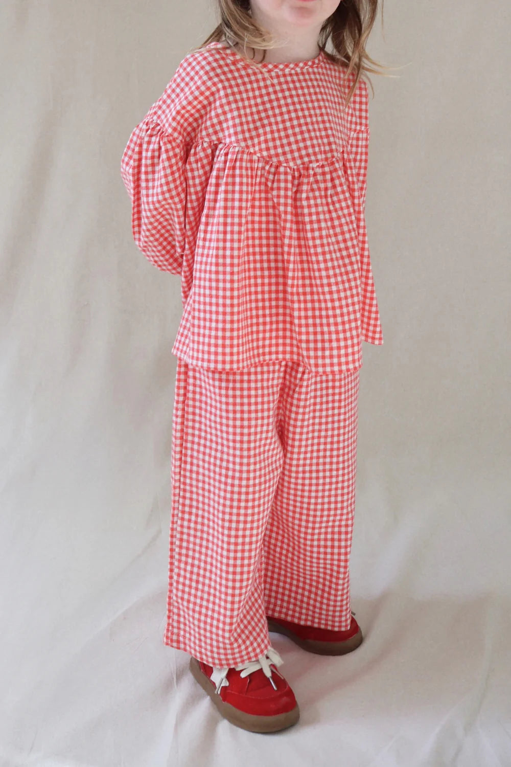 A young child stands against a light background wearing the WORN CURATIONS Gingham Bloom Blouse Chilli, styled with wide-leg pants and red shoes with white laces. The flowy long sleeves add charm, while the child's face is partly out of frame.
