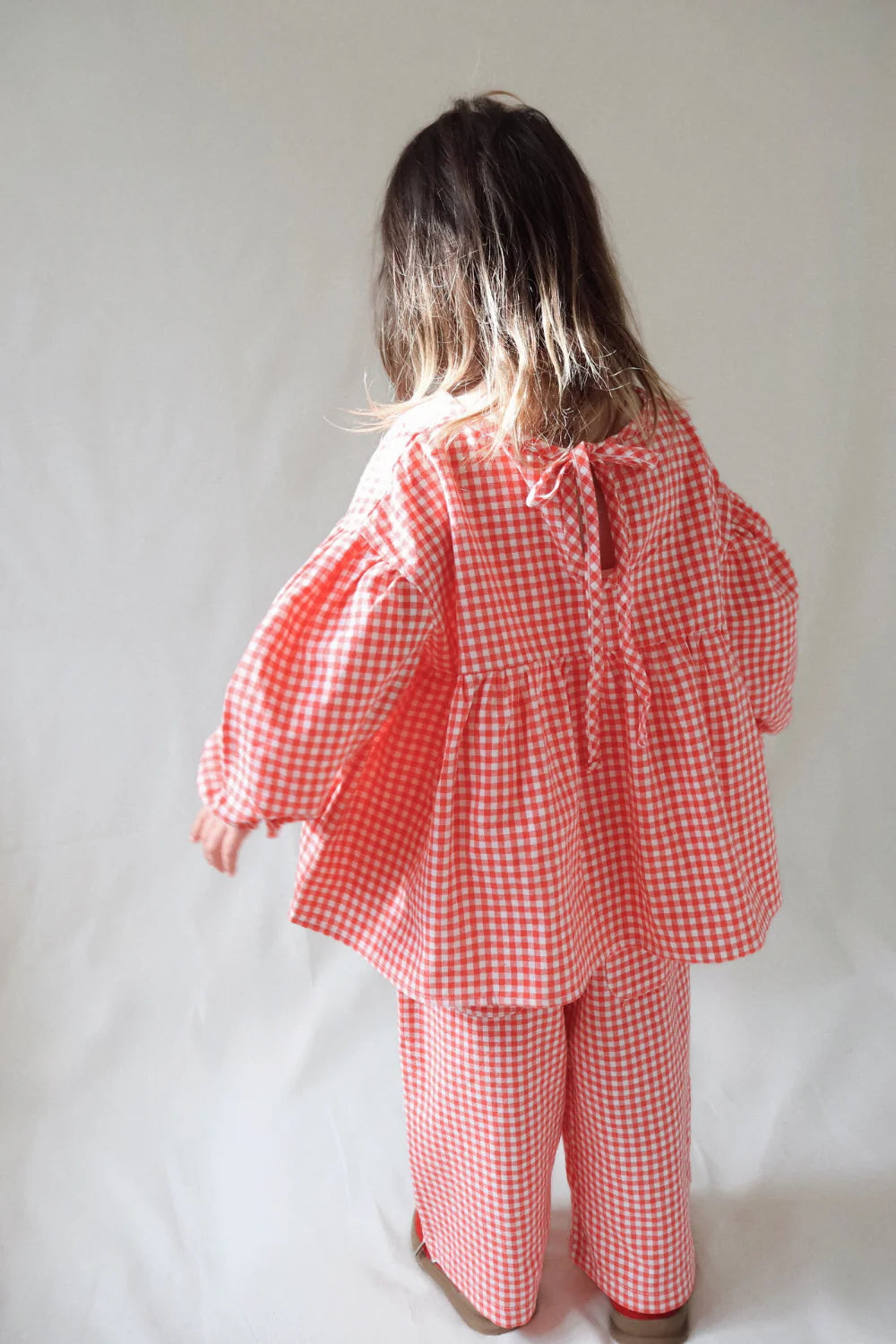 A young child with long brown hair, seen from behind, wears the WORN CURATIONS Gingham Bloom Blouse Chilli against a plain light background.