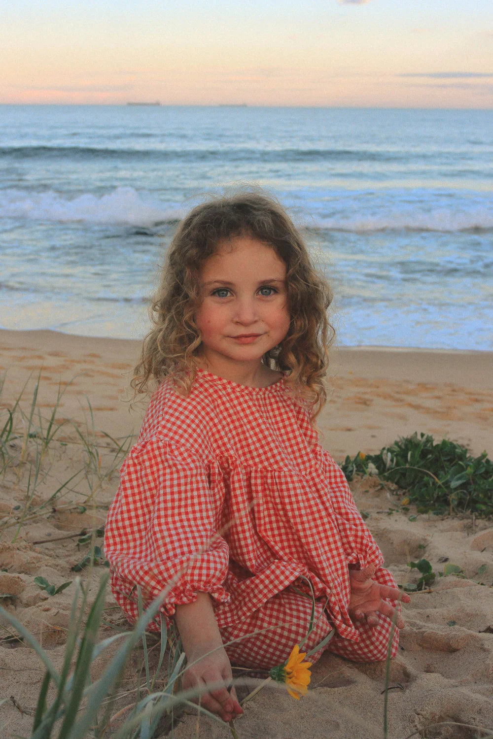 A young girl with curly hair wears the WORN CURATIONS Gingham Bloom Blouse Chilli, sitting on sandy beach grass with a yellow flower, as ocean waves and a pastel sky fill the background.
