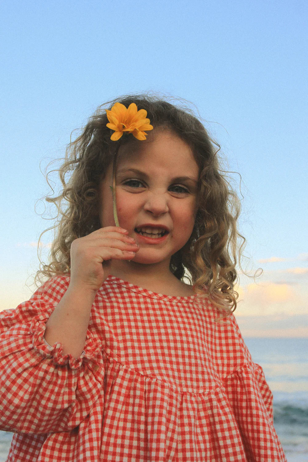 A young girl with curly hair wears the WORN CURATIONS Gingham Bloom Blouse Chilli, holding a yellow flower and smiling playfully outdoors under a blue sky with clouds.