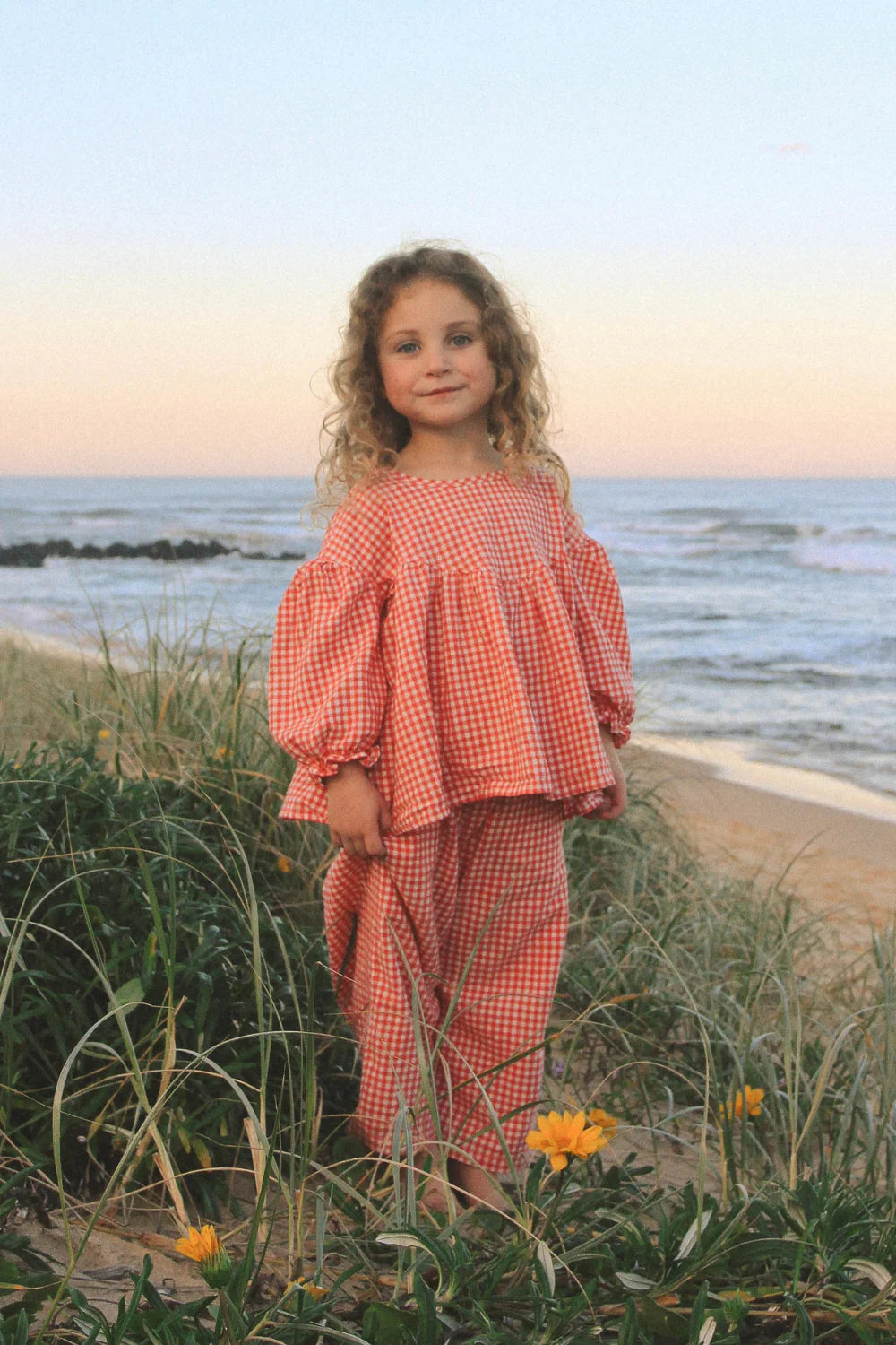 At sunset on grassy dunes, a young girl with curly hair wears the WORN CURATIONS Gingham Bloom Blouse Chilli. Yellow flowers and ocean waves add to the serene beach scene.