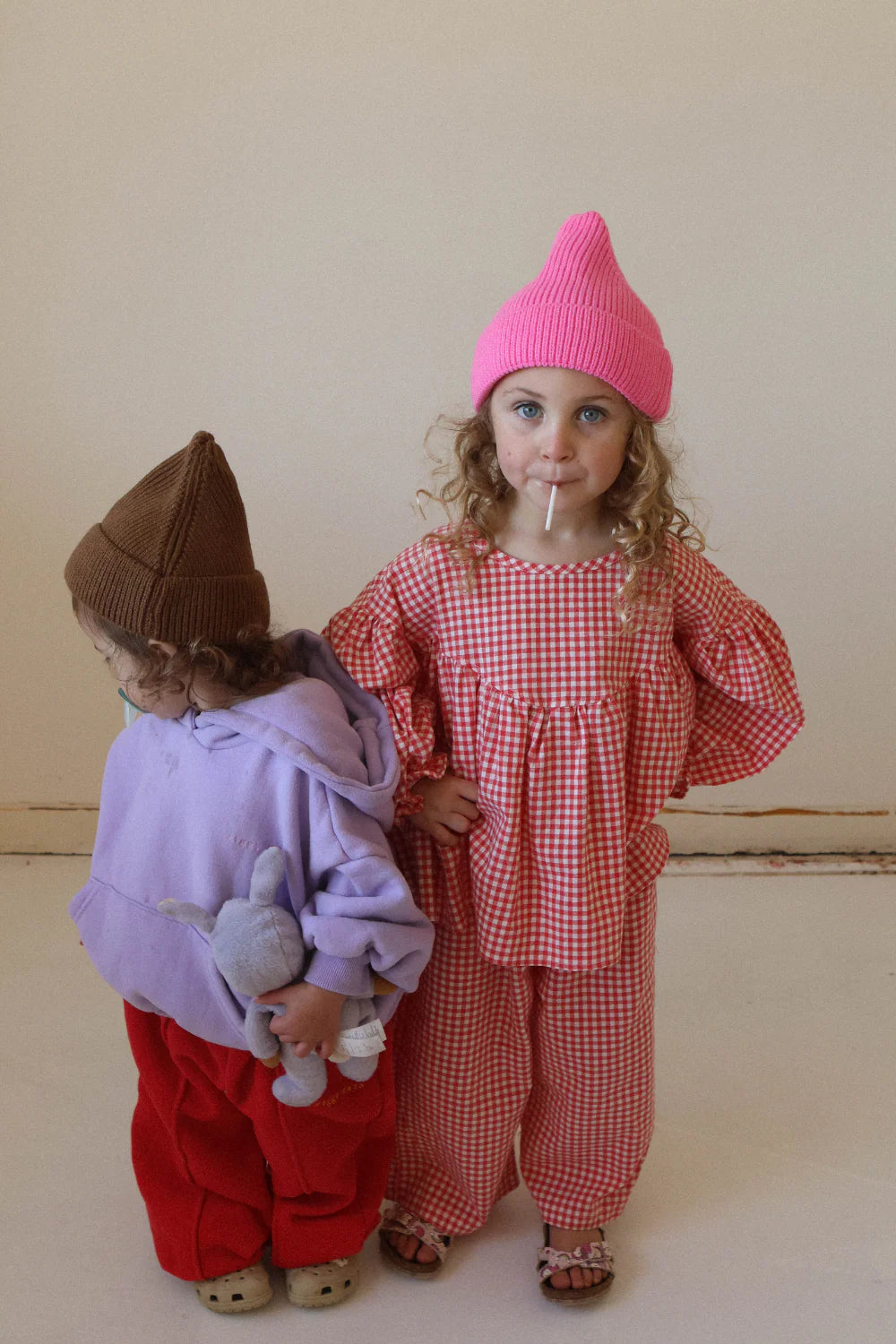 Two young children stand indoors. One wears a pink beanie and red checks with a lollipop. The other, in WORN CURATIONS’ All Day Pant Chilli, pairs them with a brown beanie, purple hoodie, and holds a stuffed bunny while looking down.