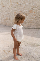 Toddler girl wearing natural romper with embroidered sleeve detail standing against a sandstone wall.
