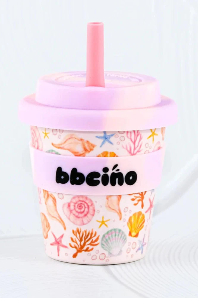 The BBcino Reusable Babycino Cup 120ml Shell, Yeah! by BBCINO features a pink lid, straw, and band labeled "bbciño." Made from sustainable bamboo, it displays vibrant sea-themed designs—shells, starfish, and coral—for an eco-friendly sip.