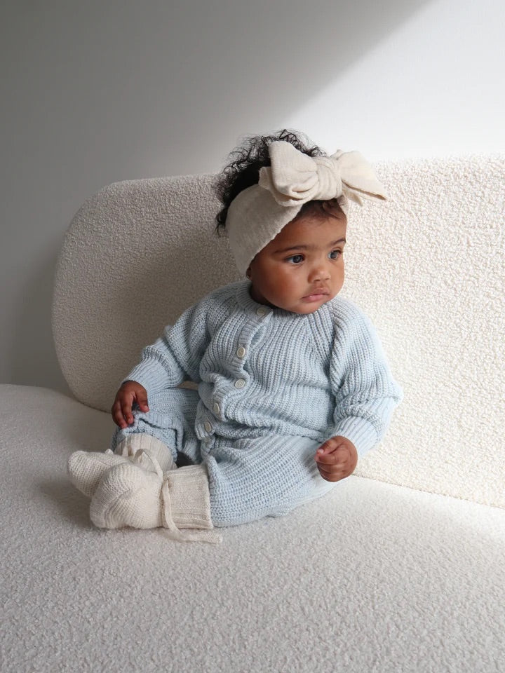 A baby sits on a textured cream sofa, wearing the ZIGGY LOU Classic Knit Romper in Aero Fleck with matching booties and a large cream bow headband, gazing thoughtfully to the side in soft natural light.