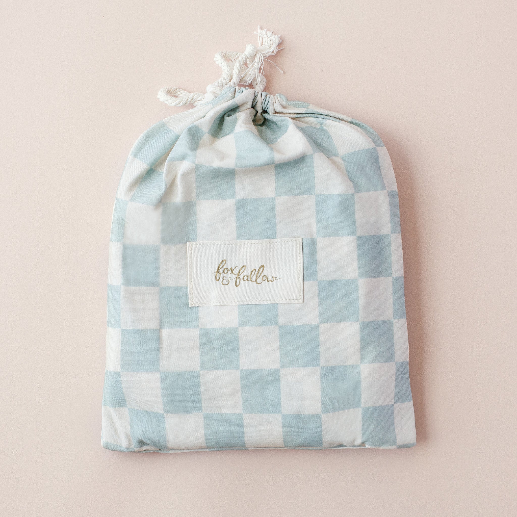 The Fox & Fallow Sage Check Organic Fitted Cot Sheet by FOX & FALLOW features a soft, GOTS certified fabric in a light blue and white check pattern, displayed against a light pink background.