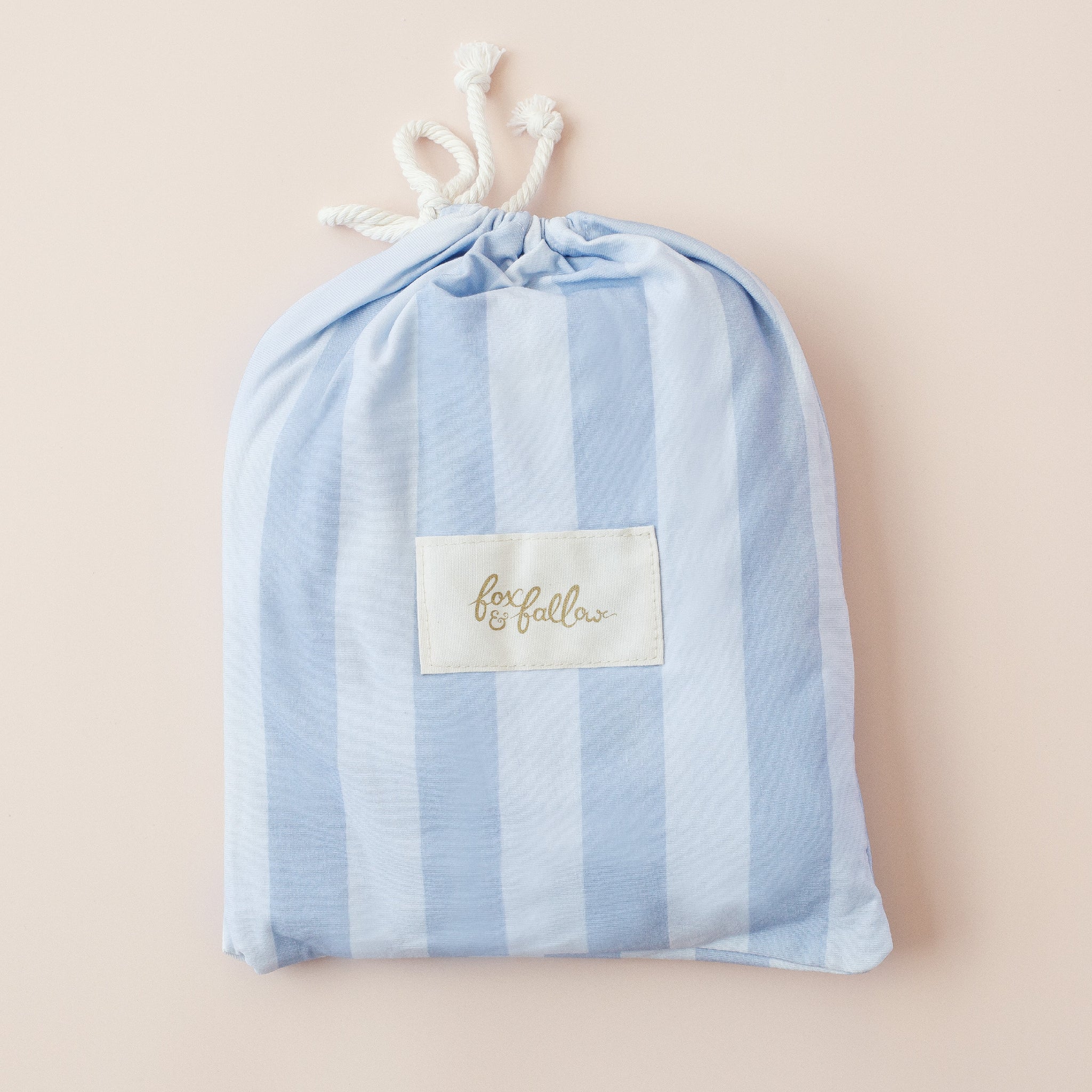 A light blue and white striped fabric drawstring bag with a "FOX & FALLOW" label on the front, perfect for storing the Fox & Fallow - Cloud Stripe Organic Fitted Cot Sheet, set against a pale pink background.