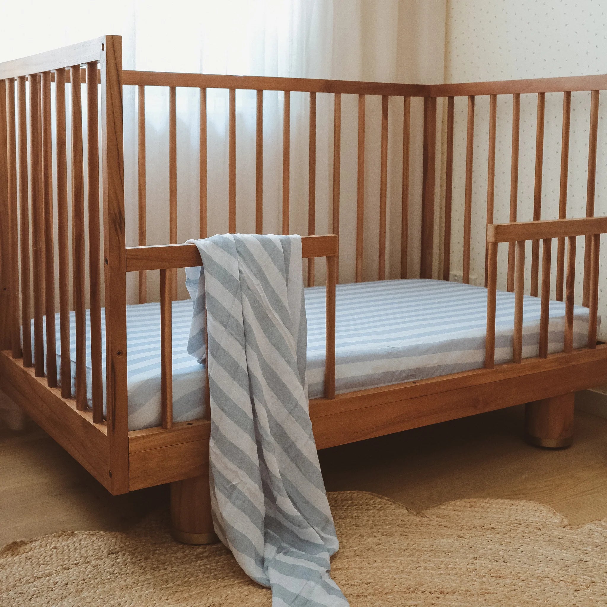 A wooden crib with vertical slats stands in a softly lit room. The FOX & FALLOW Cloud Stripe Organic Fitted Cot Sheet offers comfort, while light filters through sheer curtains onto the woven rug below.