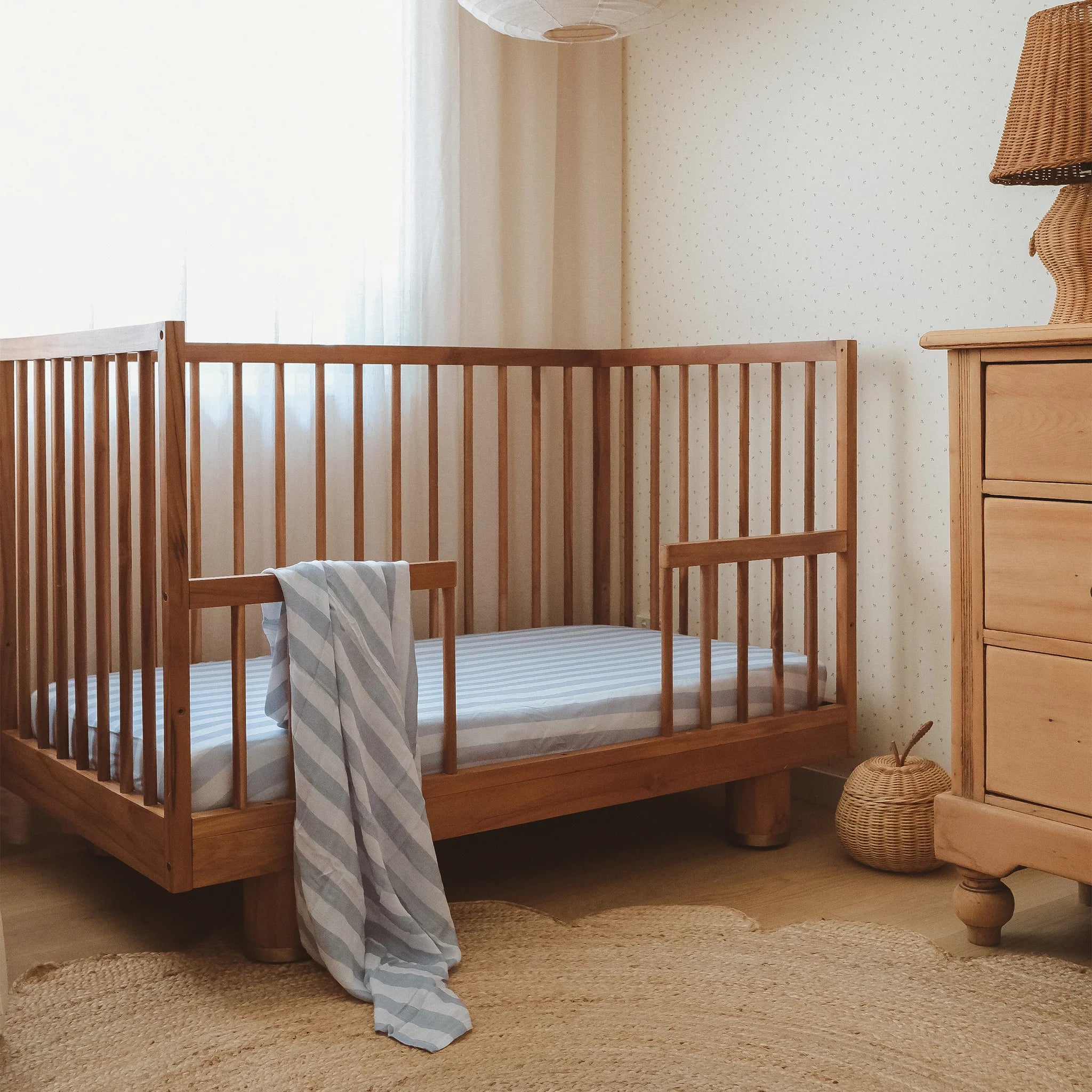 A wooden crib with a FOX & FALLOW Cloud Stripe Organic Fitted Cot Sheet and a blue-and-white striped blanket stands beside a dresser and woven basket in a softly lit, cozy nursery.