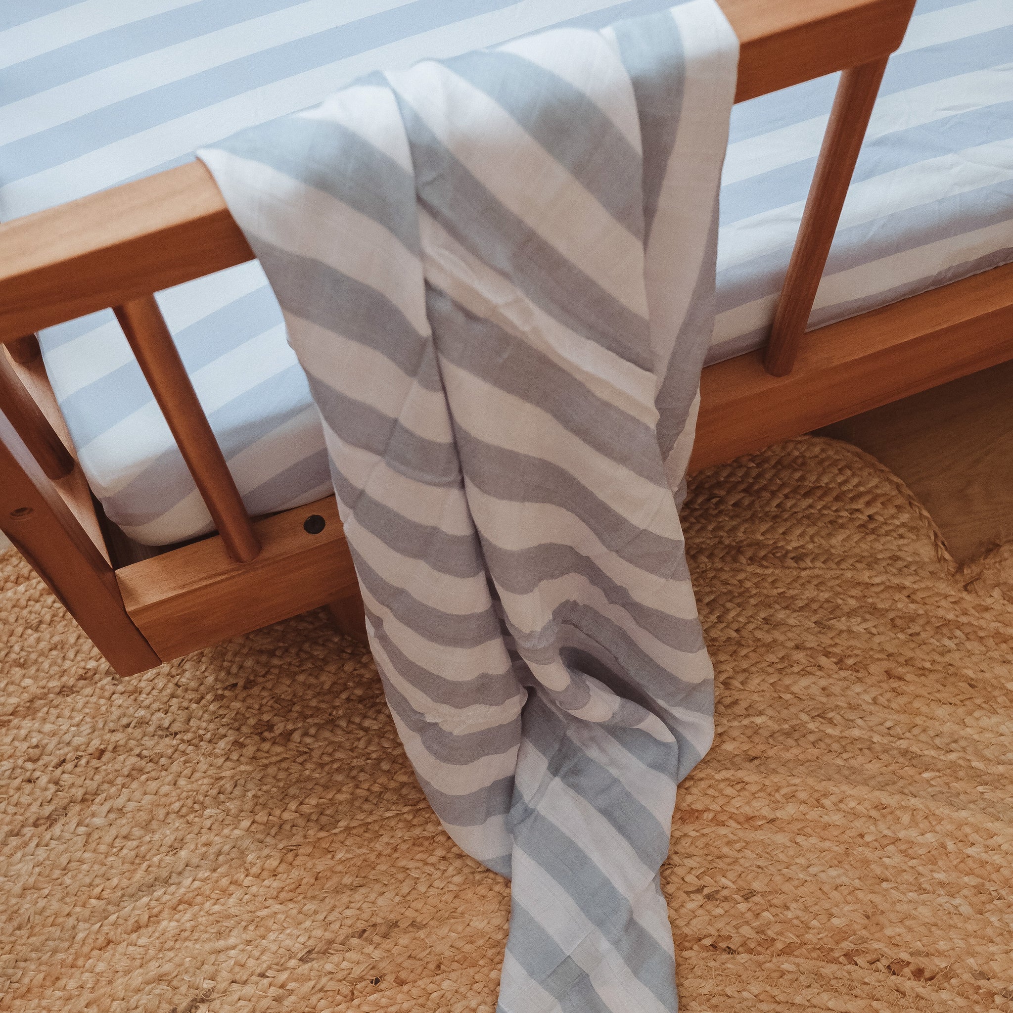 A FOX & FALLOW Cloud Stripe Organic Fitted Cot Sheet in light gray and white stripes is shown on a wooden crib, paired with a matching GOTS-certified blanket and placed atop a woven natural fiber rug.
