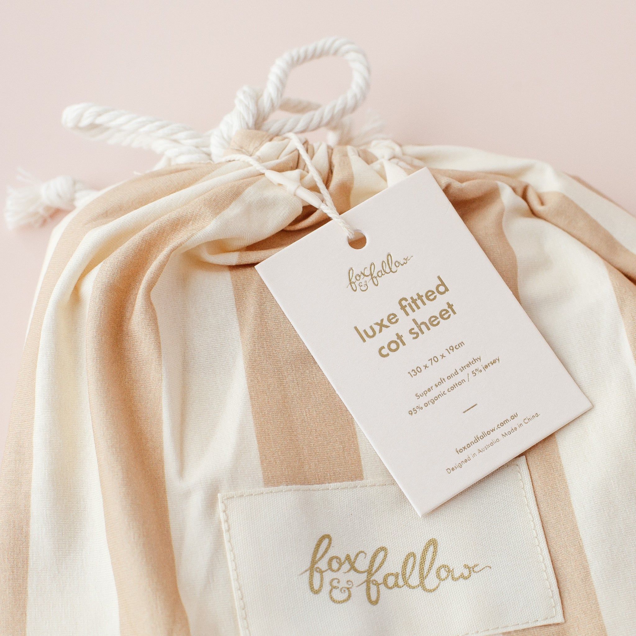 A close-up of a beige and white striped drawstring bag with "FOX & FALLOW" embroidered and a tag for the Fox & Fallow - Coffee Stripe Organic Fitted Cot Sheet, detailing size, material, and GOTS certified organic cotton.