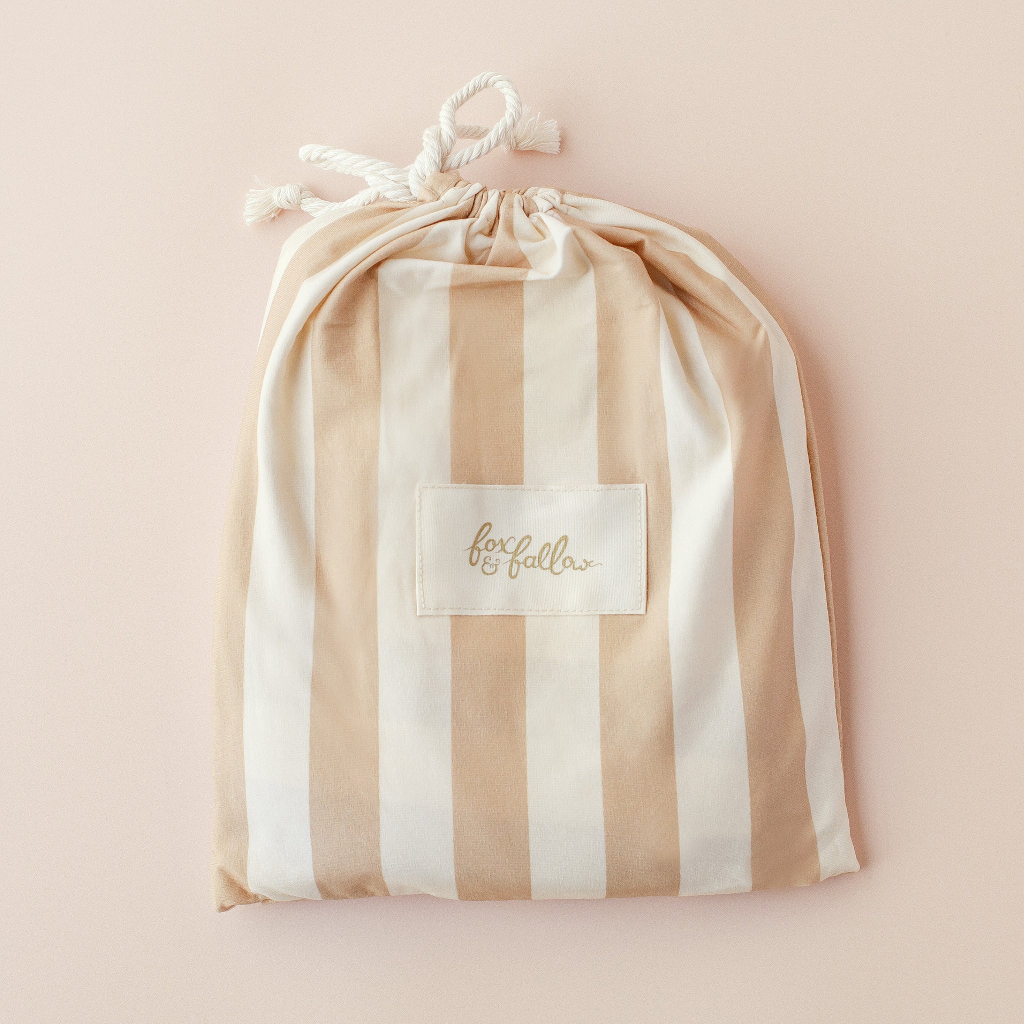 A beige and white striped drawstring bag with a white rope closure, featuring the FOX & FALLOW label, is perfect for gifting the Fox & Fallow - Coffee Stripe Organic Fitted Cot Sheet. The bag sits on a light beige background.