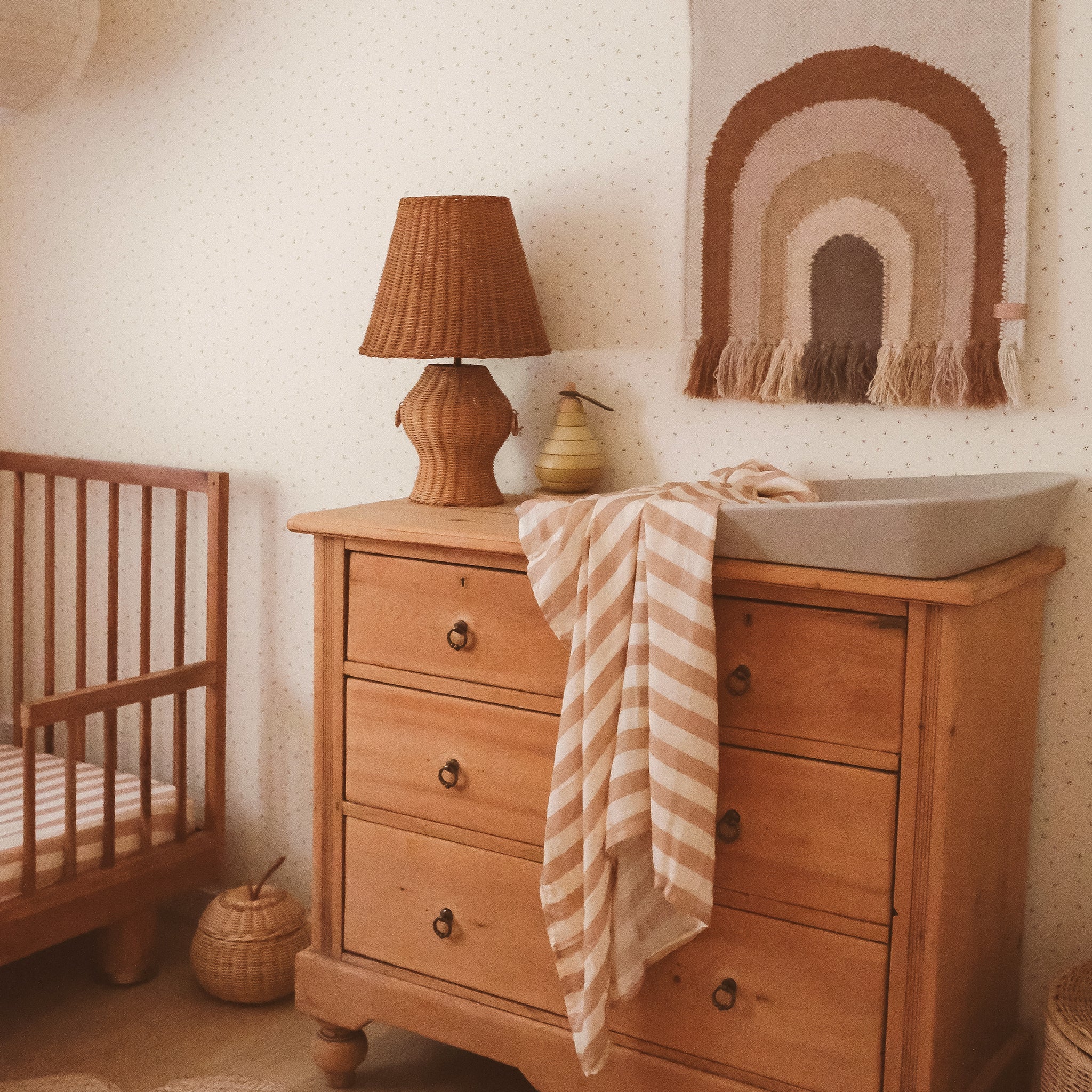 A cozy nursery featuring a wooden crib and dresser, a wicker lamp, the Fox & Fallow Coffee Stripe Organic Fitted Cot Sheet, a beige striped changing mat, a woven rainbow wall hanging, neutral decor, and soft warm lighting for a calm atmosphere.
