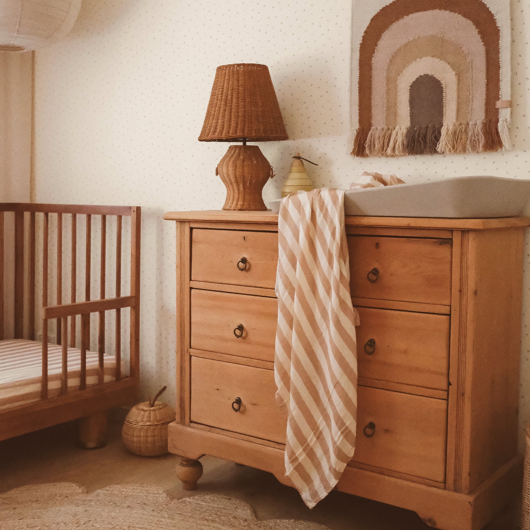 A cozy nursery features a wooden crib, dresser, and changing pad. The FOX & FALLOW Coffee Stripe Organic Fitted Cot Sheet adds warmth with its soft neutral tones, while a striped blanket and wicker lamp complete the inviting look.