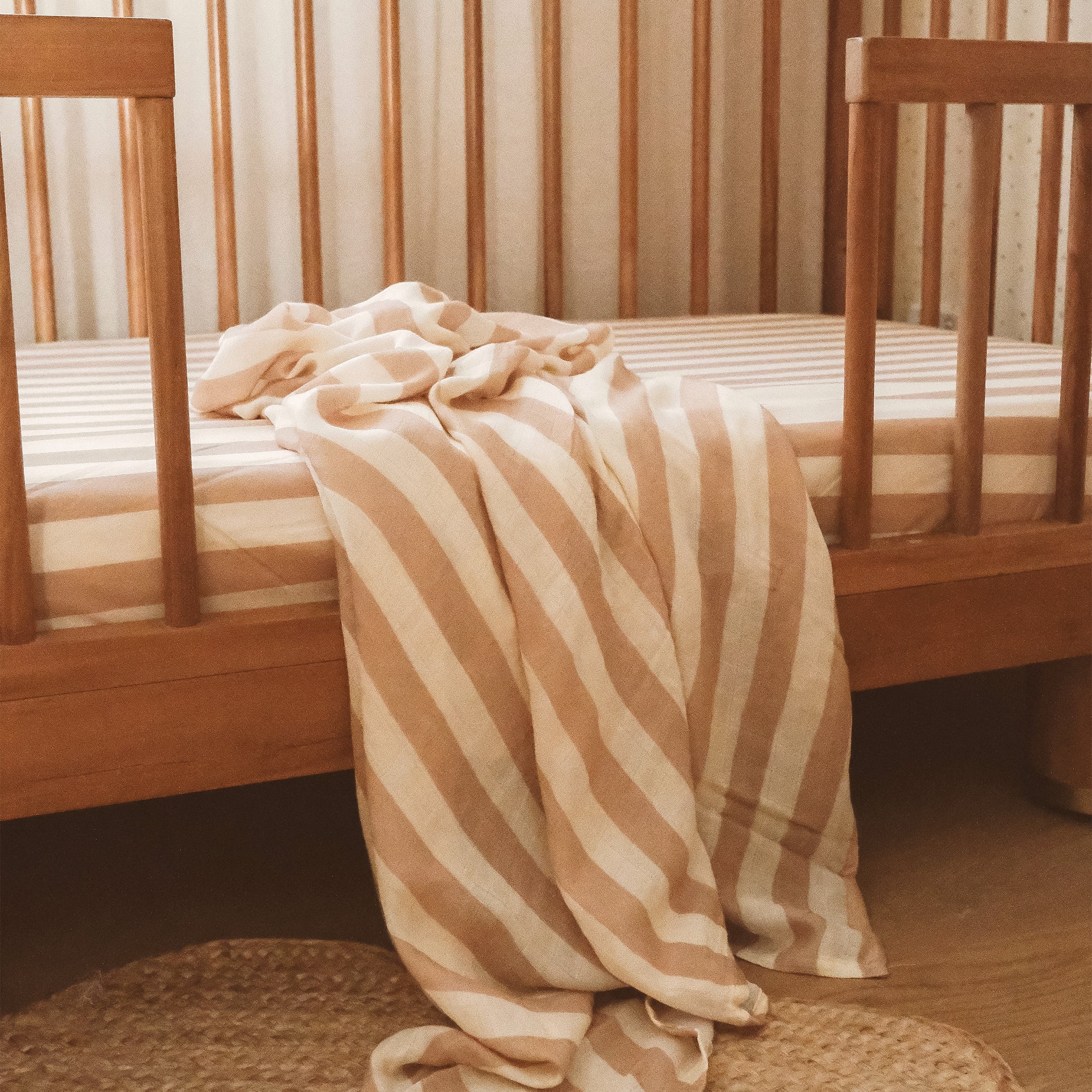 A wooden crib with a FOX & FALLOW Coffee Stripe Organic Fitted Cot Sheet and matching blanket is set on a light-colored floor, featuring beige and white stripes.