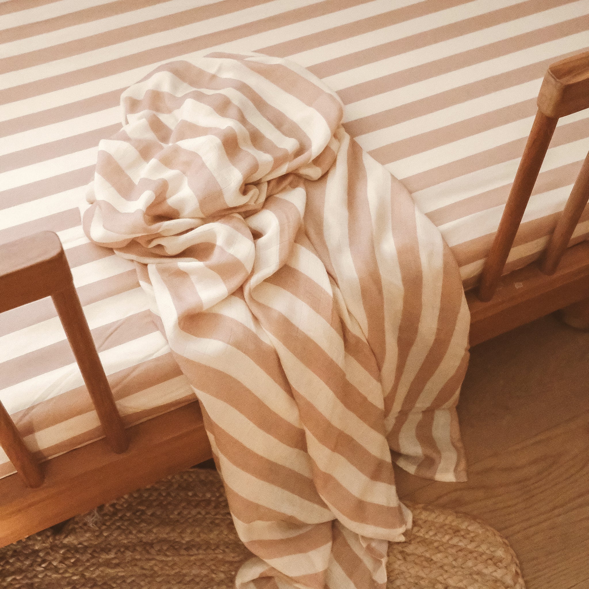 The FOX & FALLOW - Coffee Stripe Organic Fitted Cot Sheet dresses the mattress in a wooden crib, which is placed on a woven rug atop a wooden floor.