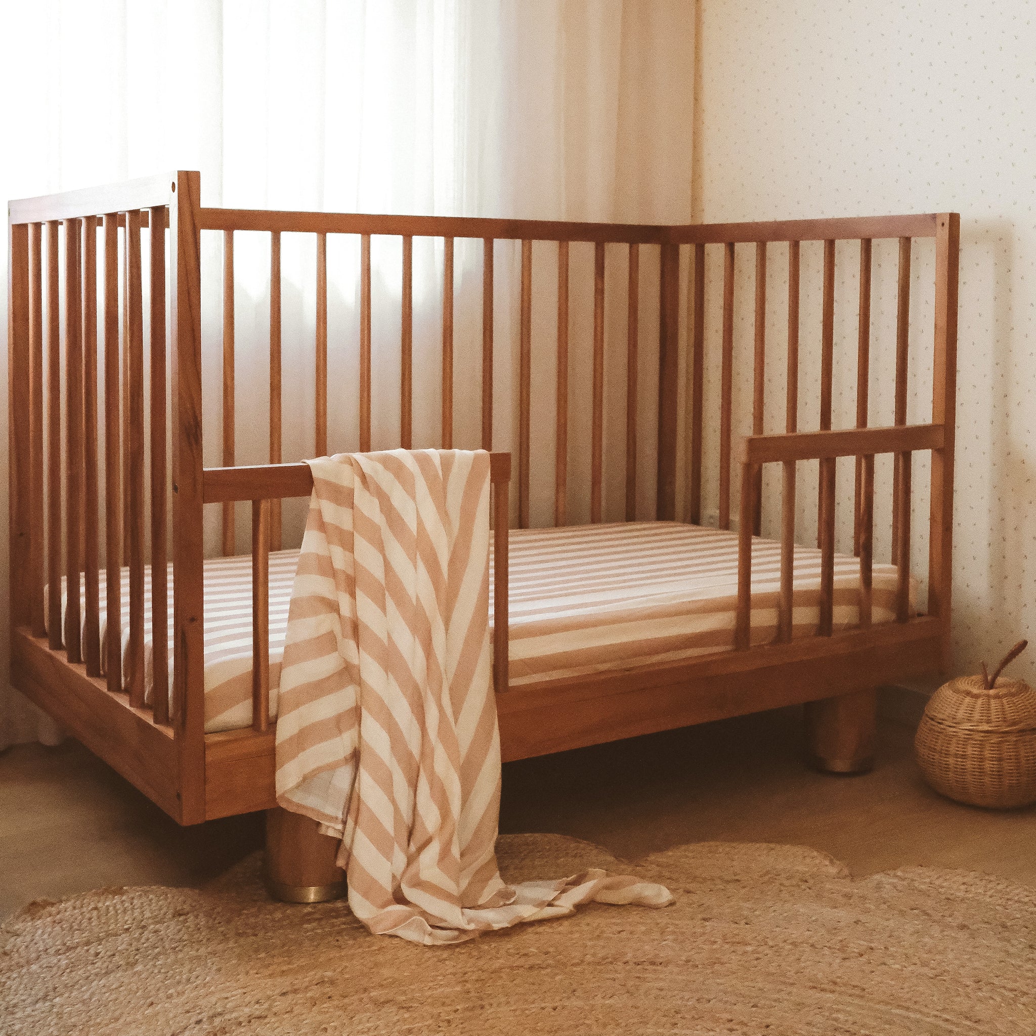 A wooden crib with vertical slats stands in a softly lit room, draped with a striped beige and white blanket. The FOX & FALLOW Coffee Stripe Organic Fitted Cot Sheet adds comfort, while a round wicker basket rests on the woven rug nearby.