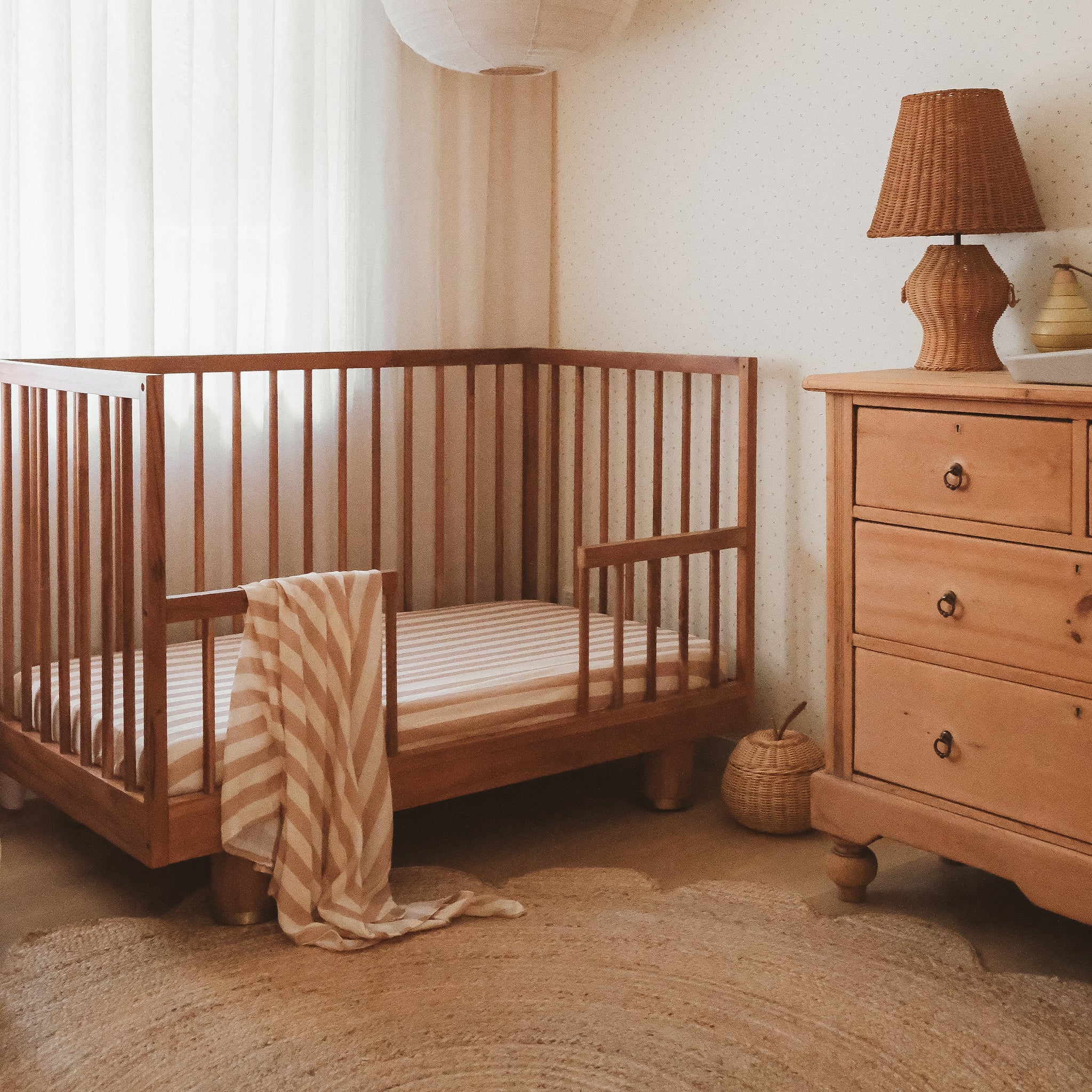 A cozy nursery with a wooden crib dressed in the FOX & FALLOW Coffee Stripe Organic Fitted Cot Sheet, a striped blanket, light curtains, a round rug, and a wooden dresser topped with a woven lamp and decor.